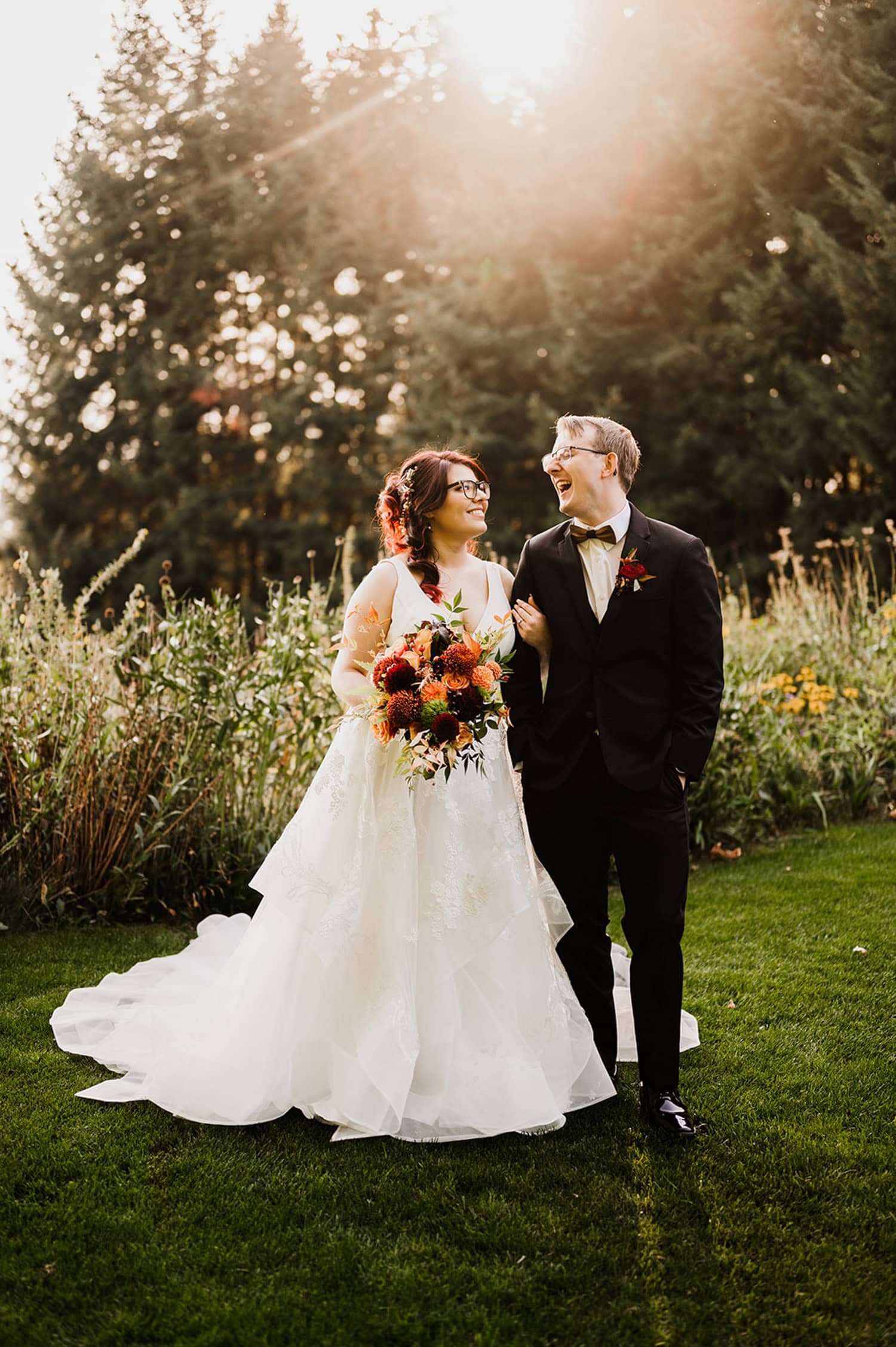 A bride and groom smiling at each other while standing on grass with a bouquet, surrounded by a forest background at Gorge Crest Vineyards with sunlight filtering through the trees.
