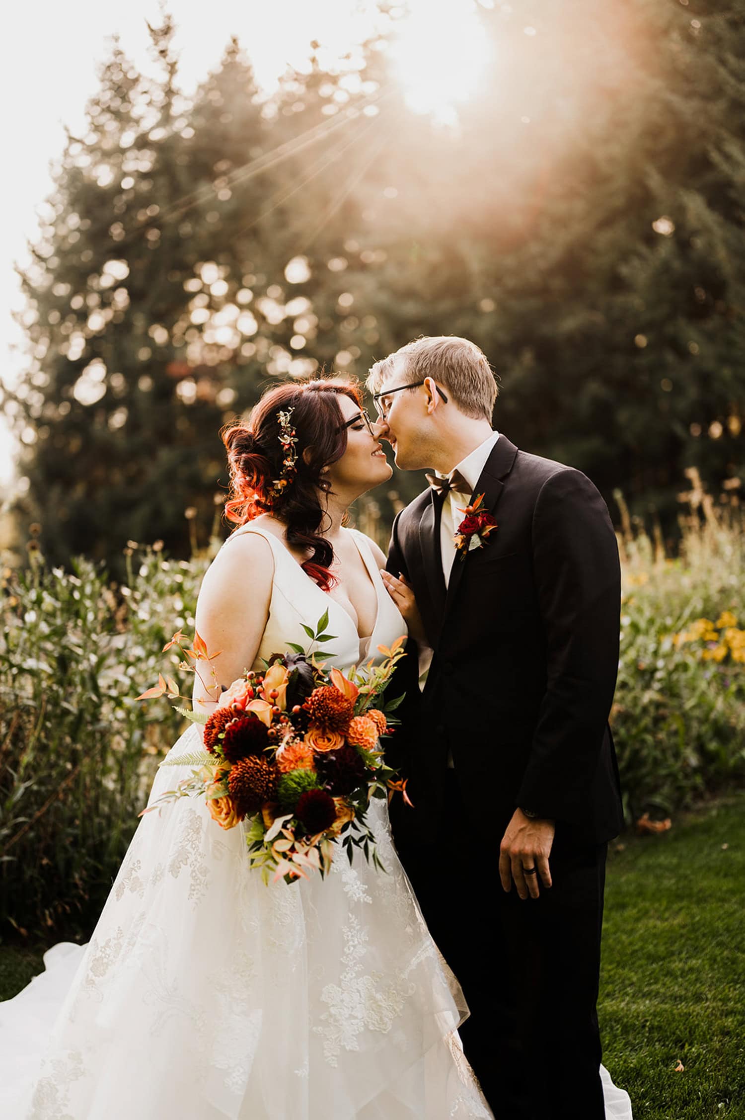 A bride and groom share a tender kiss under the sun at Gorge Crest Vineyards, holding a bouquet of vibrant orange flowers with majestic trees in the background.