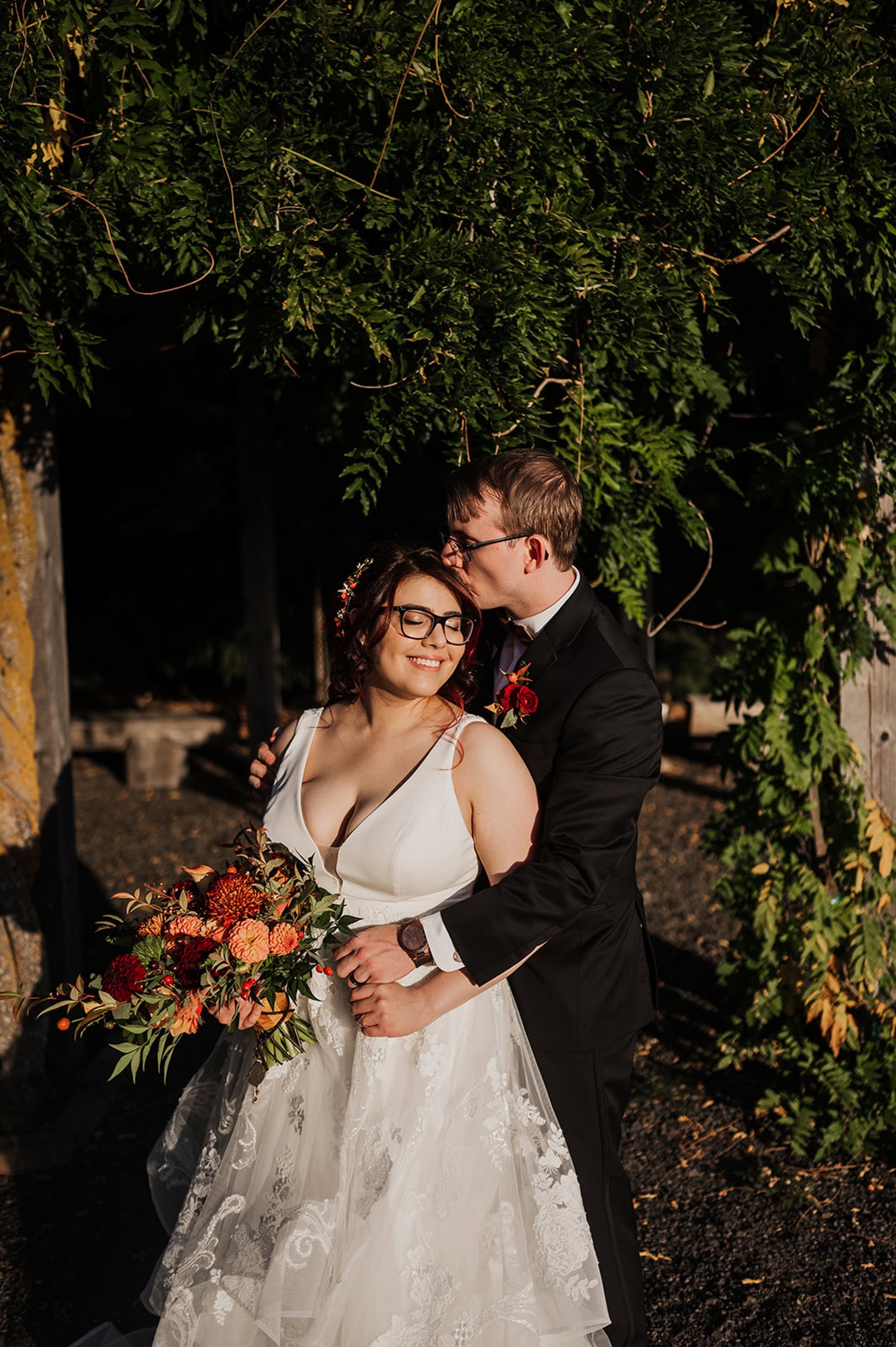 A bride and groom embrace under a leafy arch at Gorge Crest Vineyards, each holding a bouquet of flowers, smiling in the sunlight.
