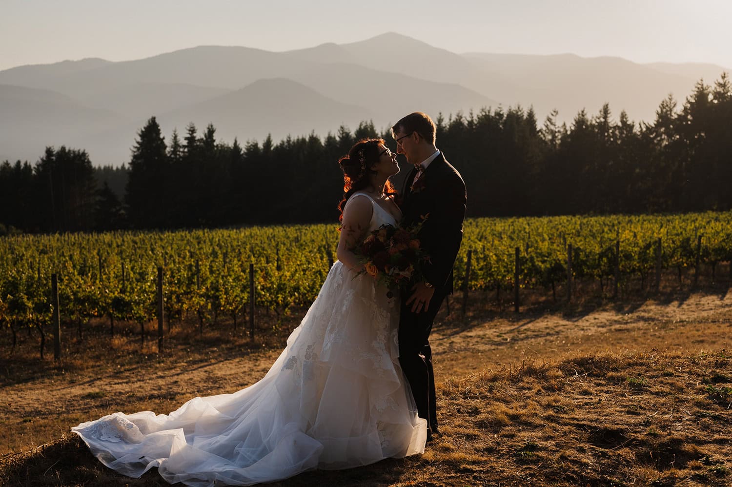 A couple in wedding attire embraces at sunset at Gorge Crest Vineyards, with mountains in the background.