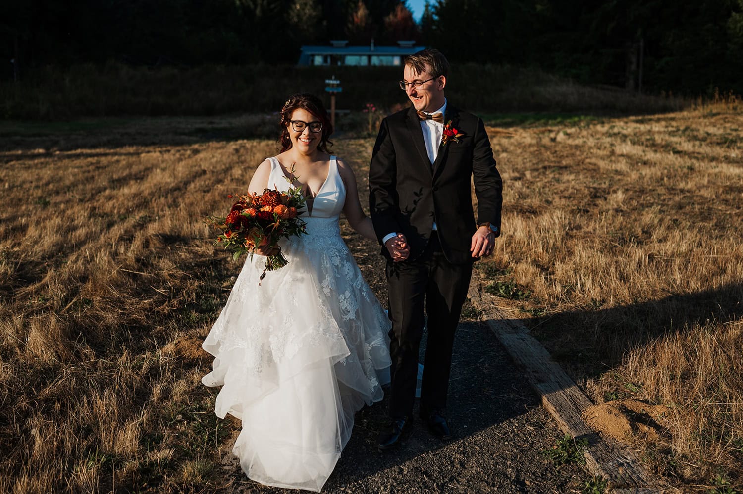 The bride and groom hold hands while walking outside on a sunny day at Gorge Crest Vineyards. The bride carries a bouquet of vibrant orange flowers.