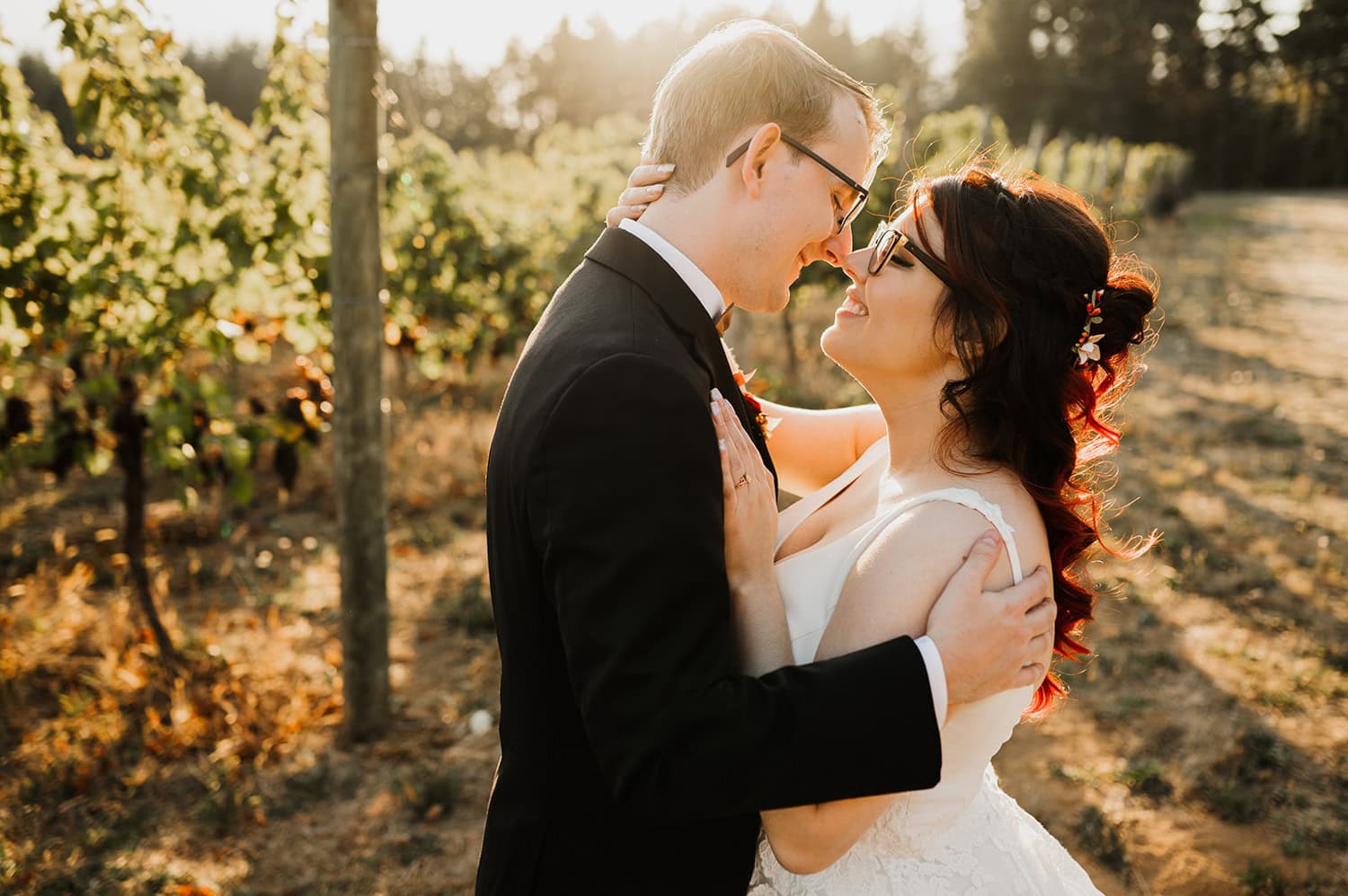 A couple in wedding attire is smiling and embracing outdoors at sunset in Gorge Crest Vineyards.