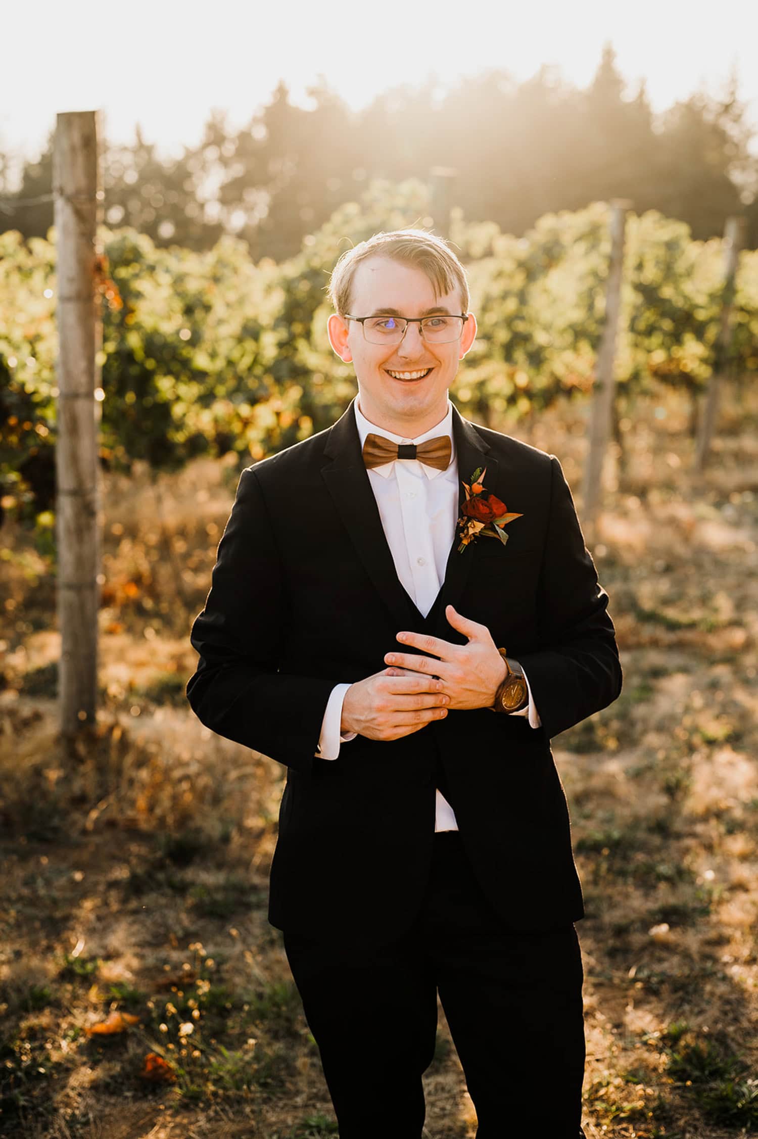 A groom in a black suit and bow tie stands with hands clasped, smiling at Gorge Crest Vineyards during sunset.