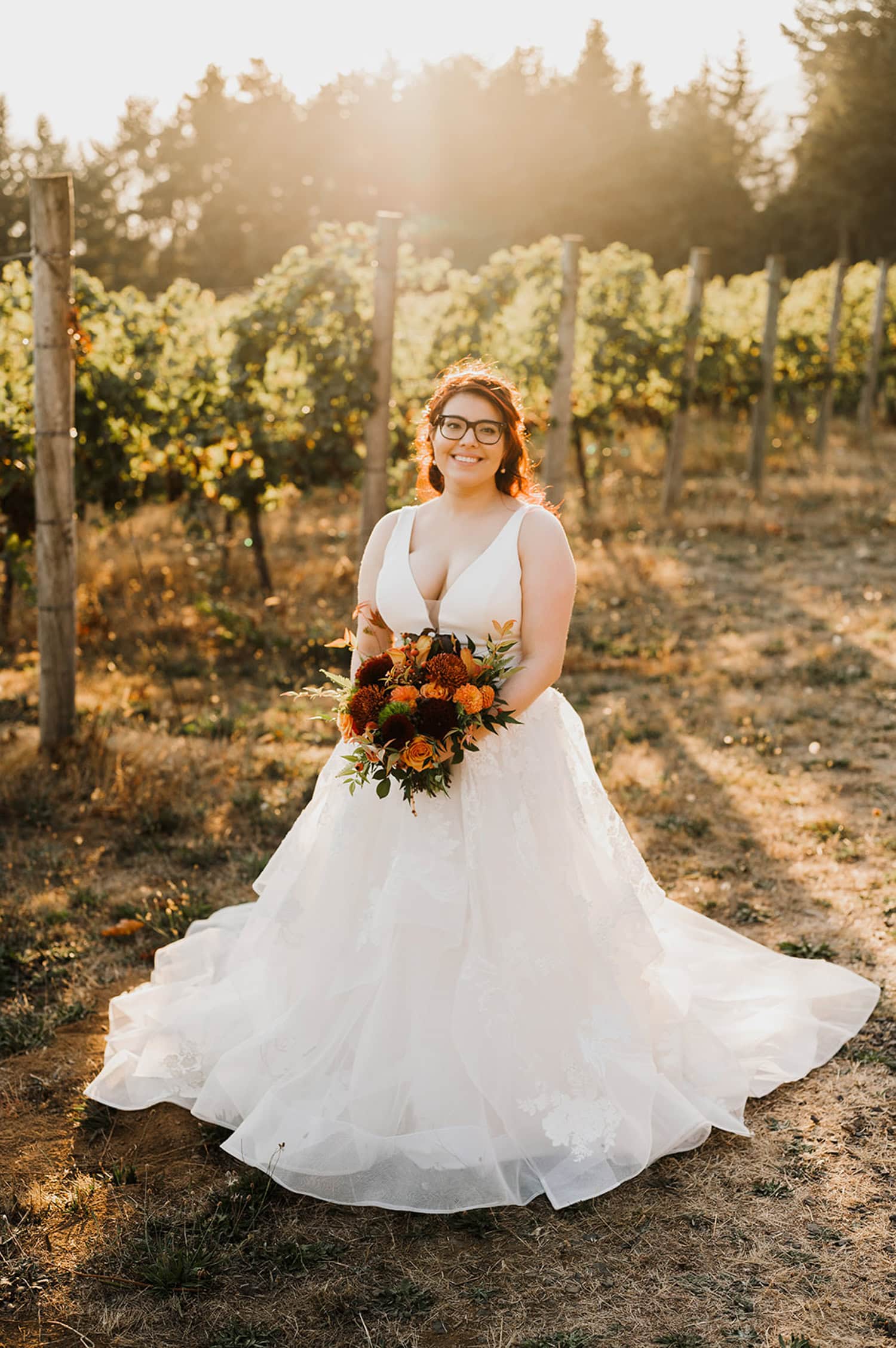 A bride in a white gown holds a bouquet of red and orange flowers, standing gracefully at sunset in the scenic Gorge Crest Vineyards.