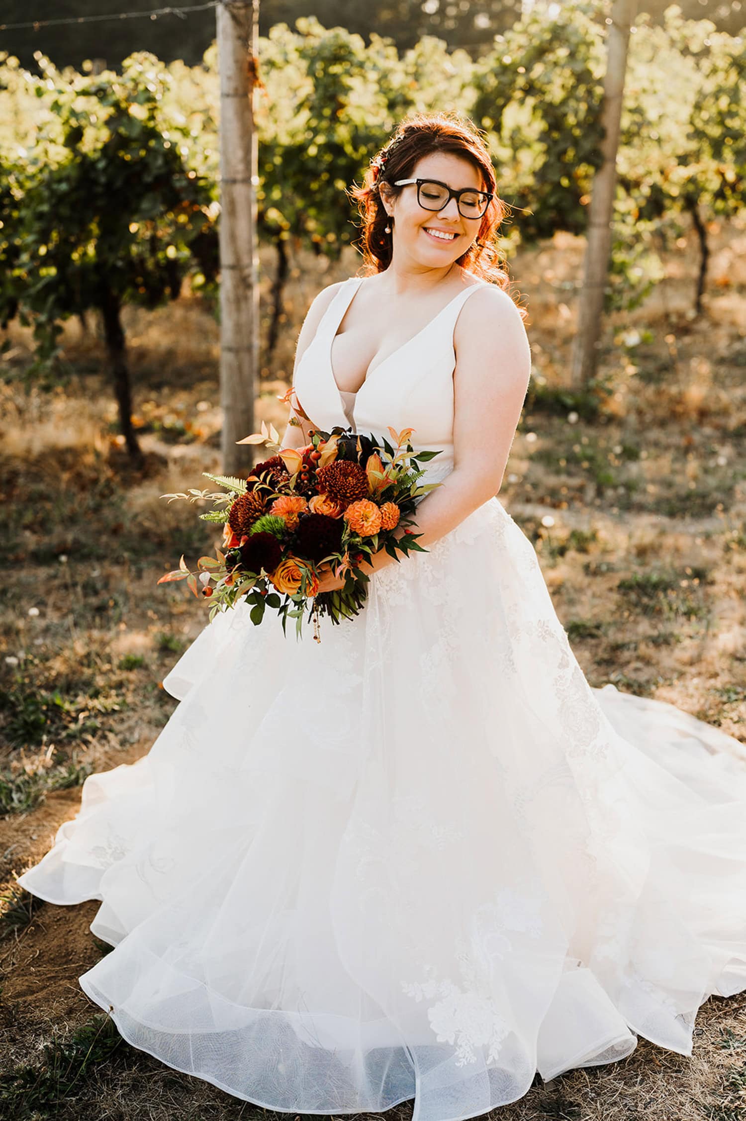 A bride in a white gown holds an orange bouquet, standing in the sunlight at Gorge Crest Vineyards.