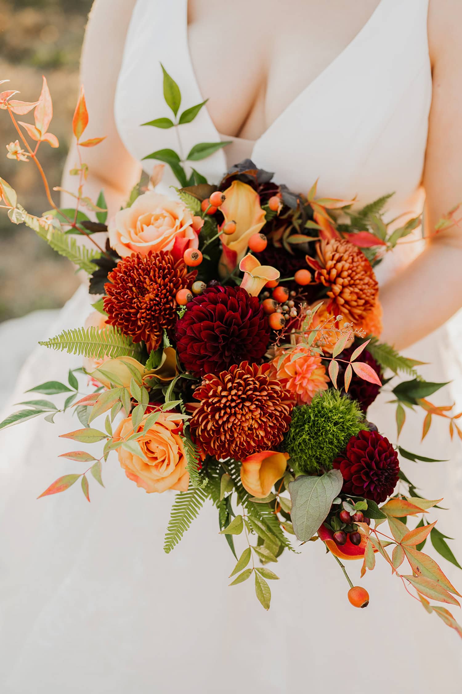 A bride holding a bouquet with red, orange, and peach flowers with green leaves, standing in front of the scenic views at Gorge Crest Vineyards.