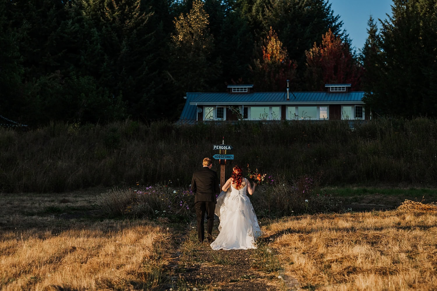 A bride and groom walk hand in hand along a path at Gorge Crest Vineyards, surrounded by lush greenery and sprawling vineyards under a clear blue sky. Their attire contrasts beautifully with the natural landscape, creating a harmonious blend of elegance and nature.
