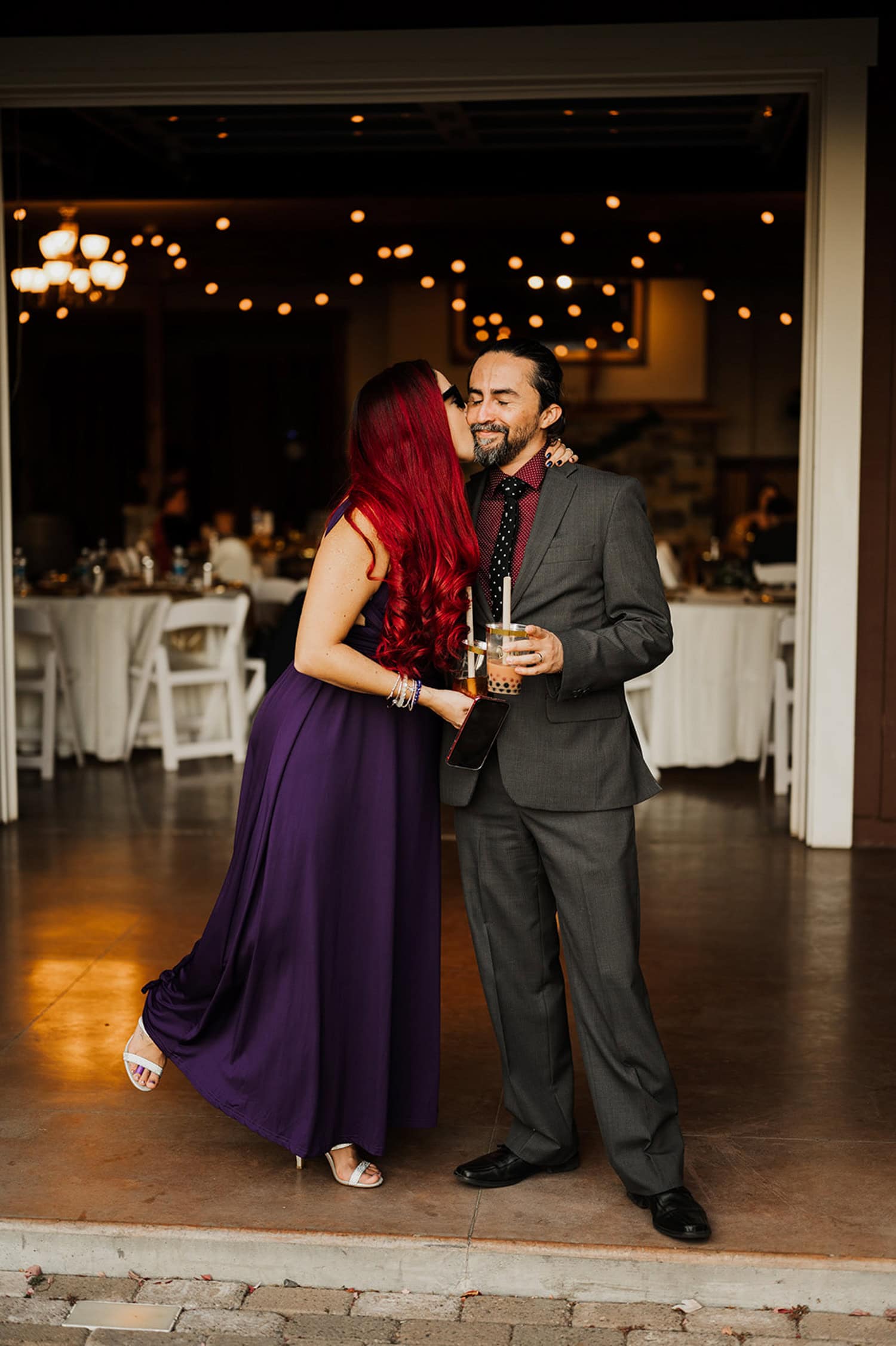 At a Gorge Crest Vineyards wedding, a woman in a purple dress and a man in a suit share a moment beneath twinkling string lights, surrounded by the festive atmosphere of the gathering.