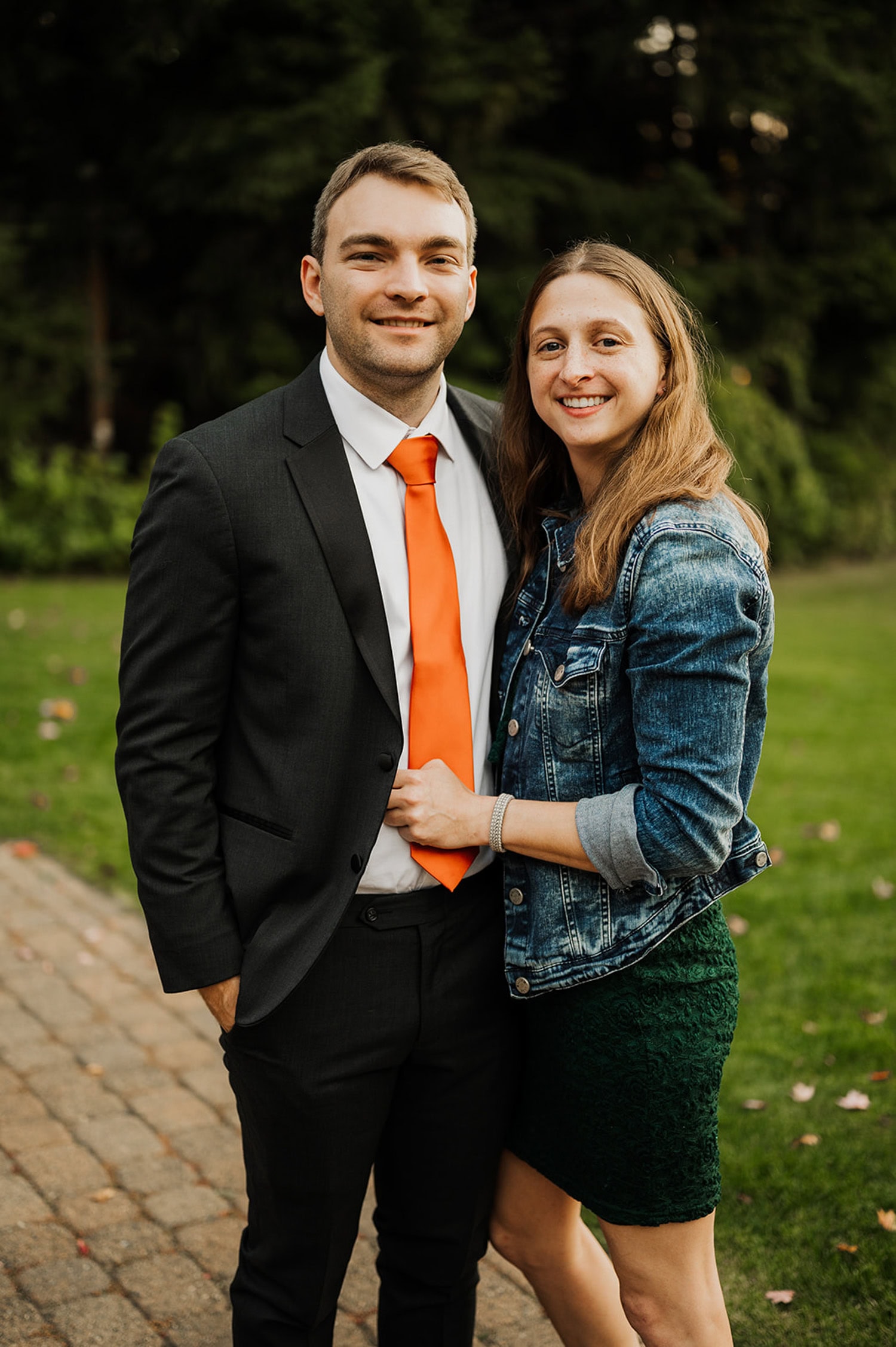 A couple poses outdoors at Gorge Crest Vineyards; the man wears a suit with an orange tie, and the woman is in a denim jacket and green dress. Lush vineyard landscape in the background.