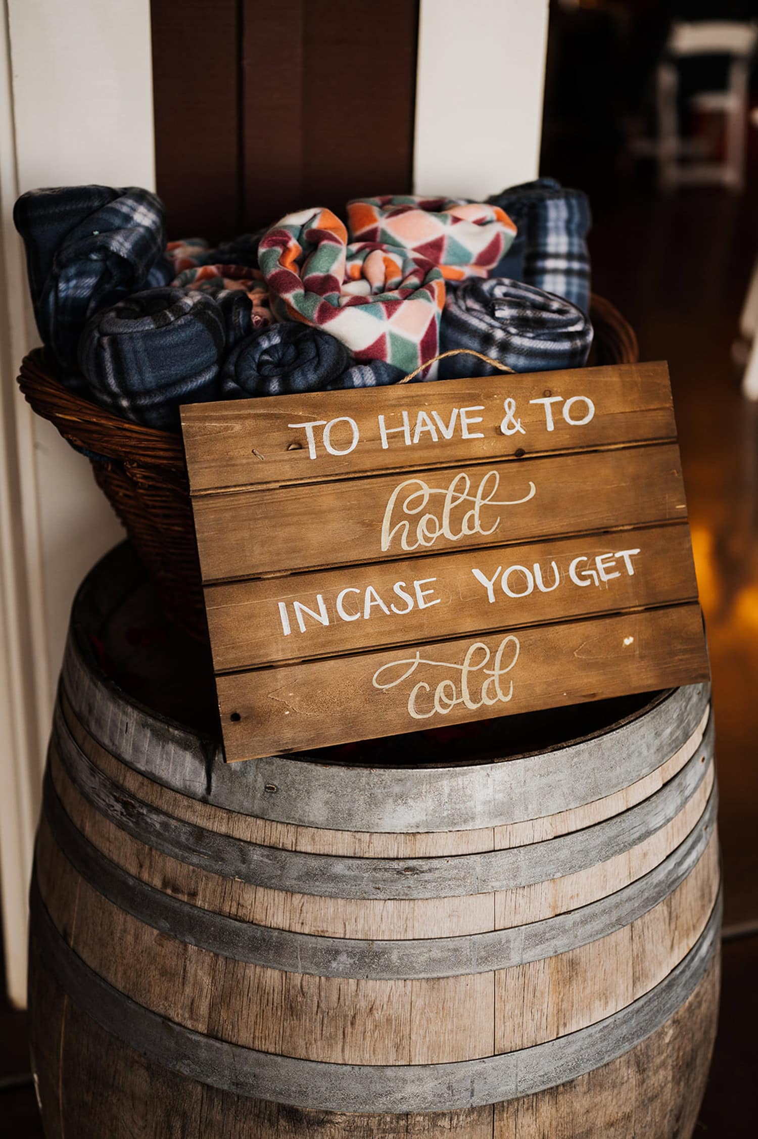 A cozy basket filled with rolled blankets sits on a barrel next to a wooden sign that reads, To have & to hold in case you get cold. The setup is part of a Gorge Crest Vineyards wedding.