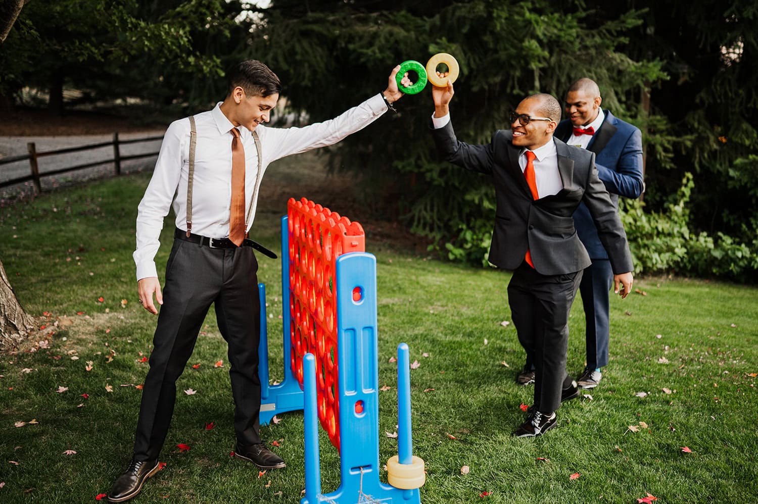 Three men in suits play Connect Four outdoors at Gorge Crest Vineyards, smiling and interacting with large game pieces at a wedding.