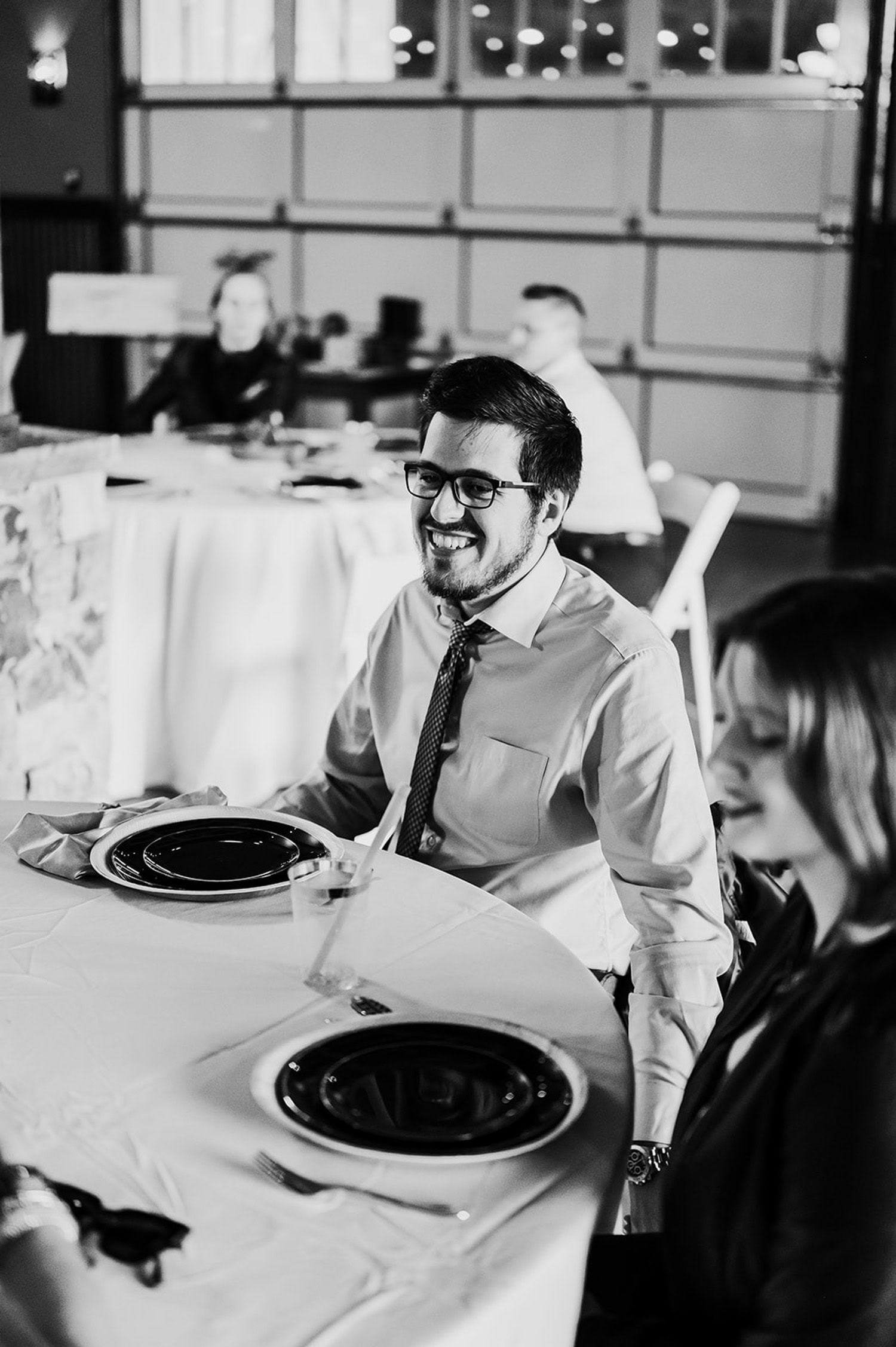 A man smiles at a table with black plates and a glass, set against the backdrop of Gorge Crest Vineyards; blurred guests are mingling in the background.