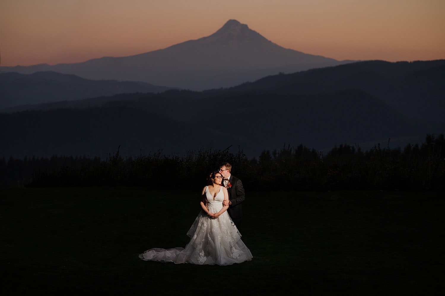 The bride and groom embrace at dusk, silhouetted against a mountain backdrop with a dimly lit sky at Gorge Crest Vineyards.