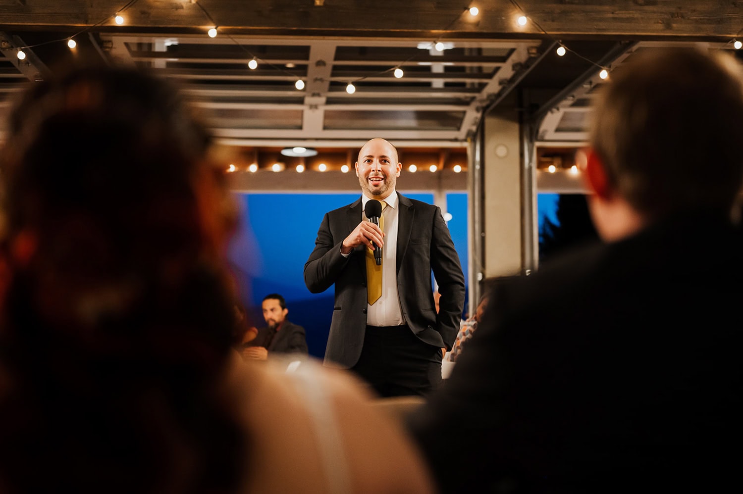 At a Gorge Crest Vineyards wedding, an individual in a suit speaks passionately into a microphone, illuminated by the warm glow of string lights. The audience is blurred in the foreground, listening intently.