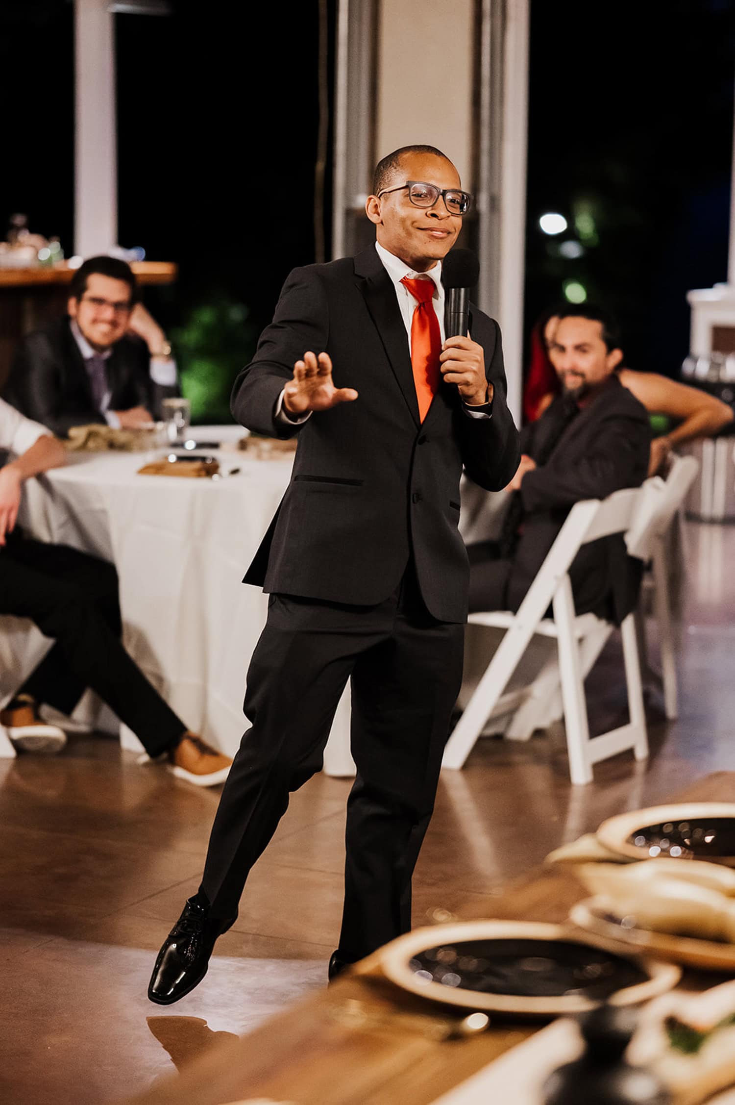 A person in a suit and red tie speaks passionately into a microphone at an event, gesturing animatedly. Seated in the background is an attentive audience, with a scenic view suggesting an outdoor venue.