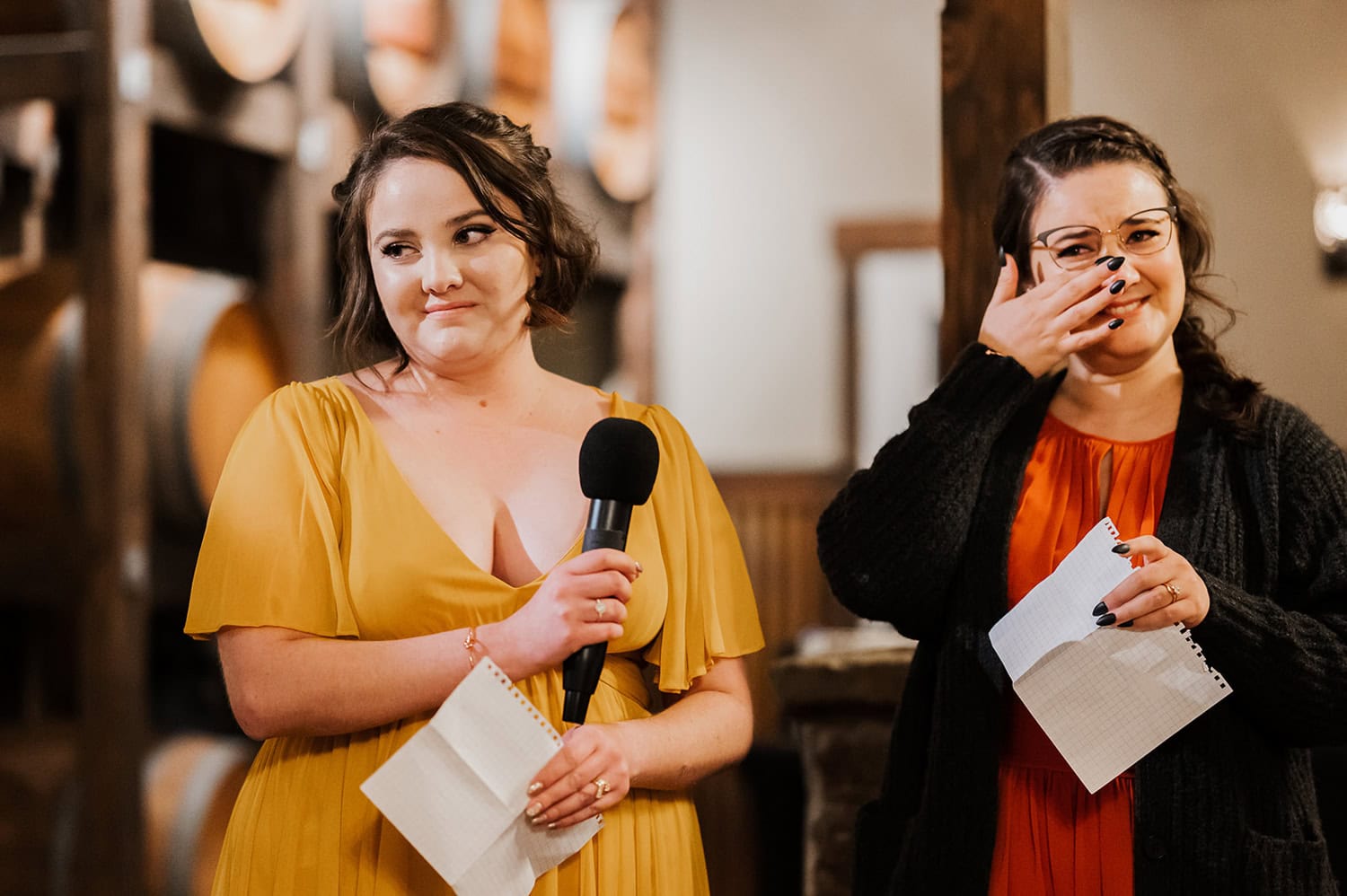 Two women stand close together at a Gorge Crest Vineyards wedding; one is holding a microphone, and the other is wiping away a tear in a warmly lit room.