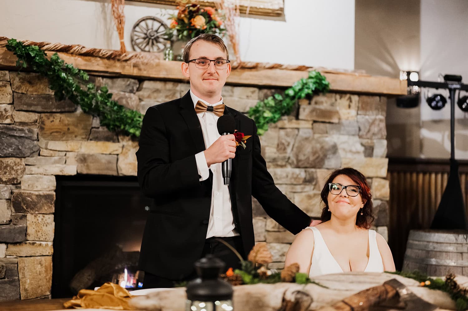 A person in a suit holds a microphone while standing next to a seated person in a dress, surrounded by lush greenery at Gorge Crest Vineyards during a wedding ceremony.