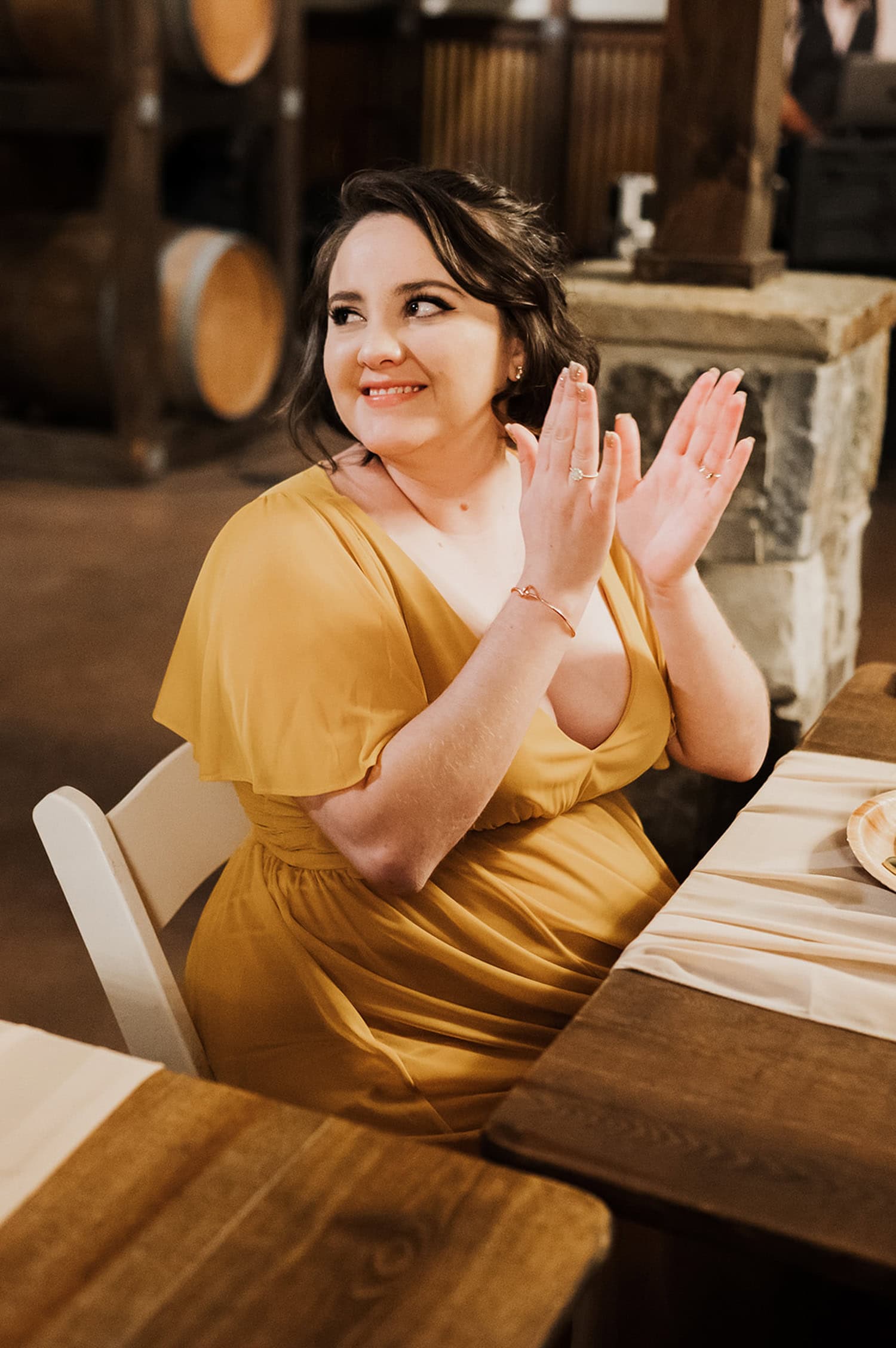 A woman in a yellow dress is seated at a wooden table indoors at Gorge Crest Vineyards, clapping and smiling with delight.