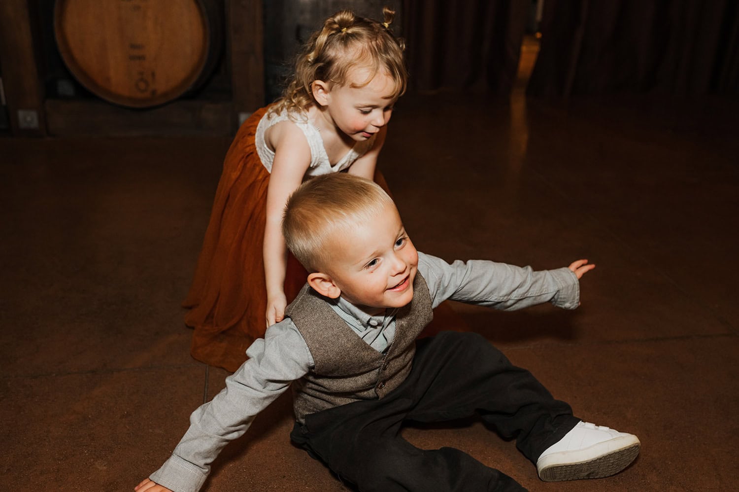 Two children play indoors; a boy sits on the floor smiling, while a girl in a dress stands behind him.