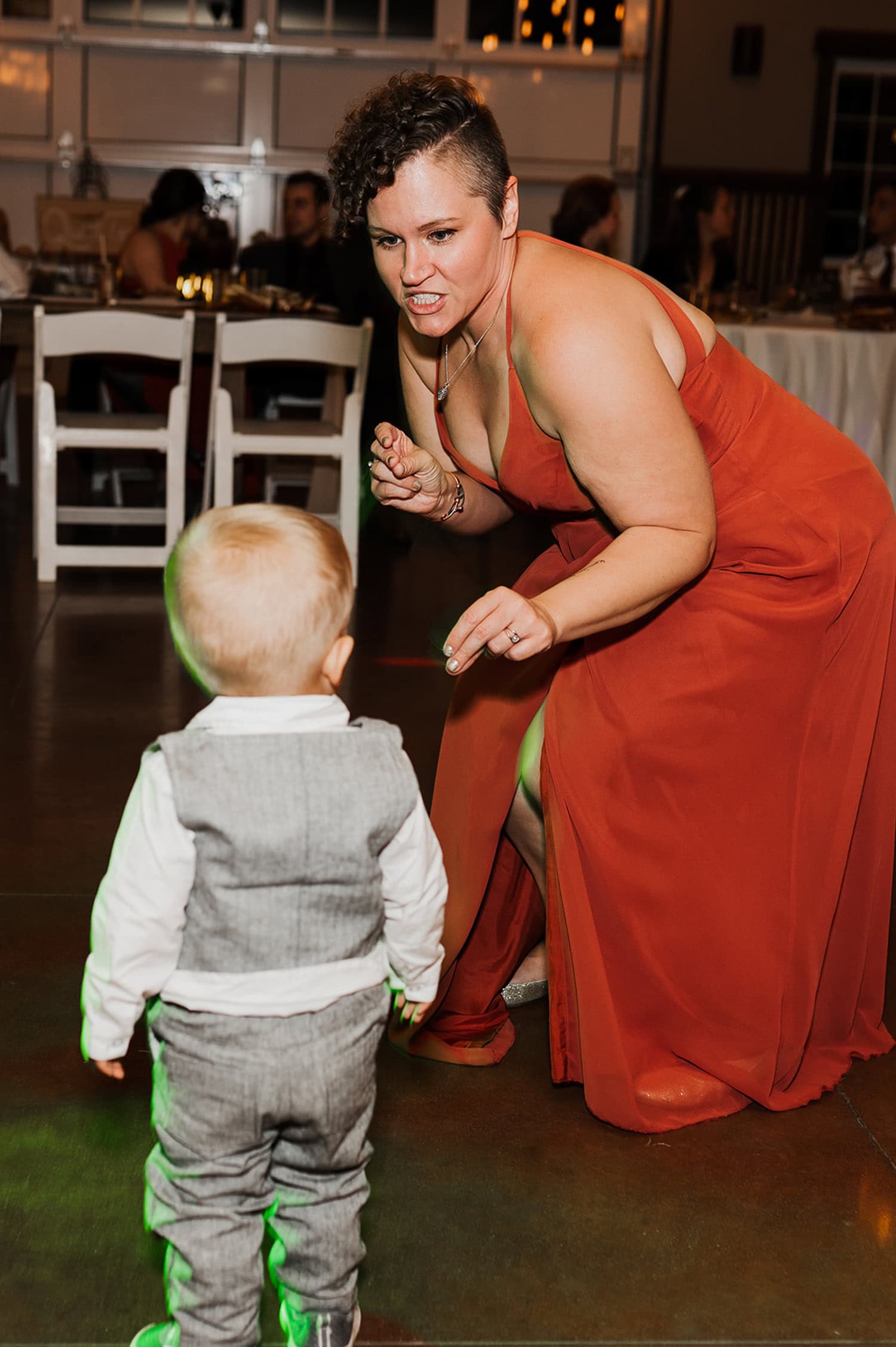 A woman in a red dress joyfully dances with a toddler in a gray suit at an indoor event, both laughing and appearing full of energy.