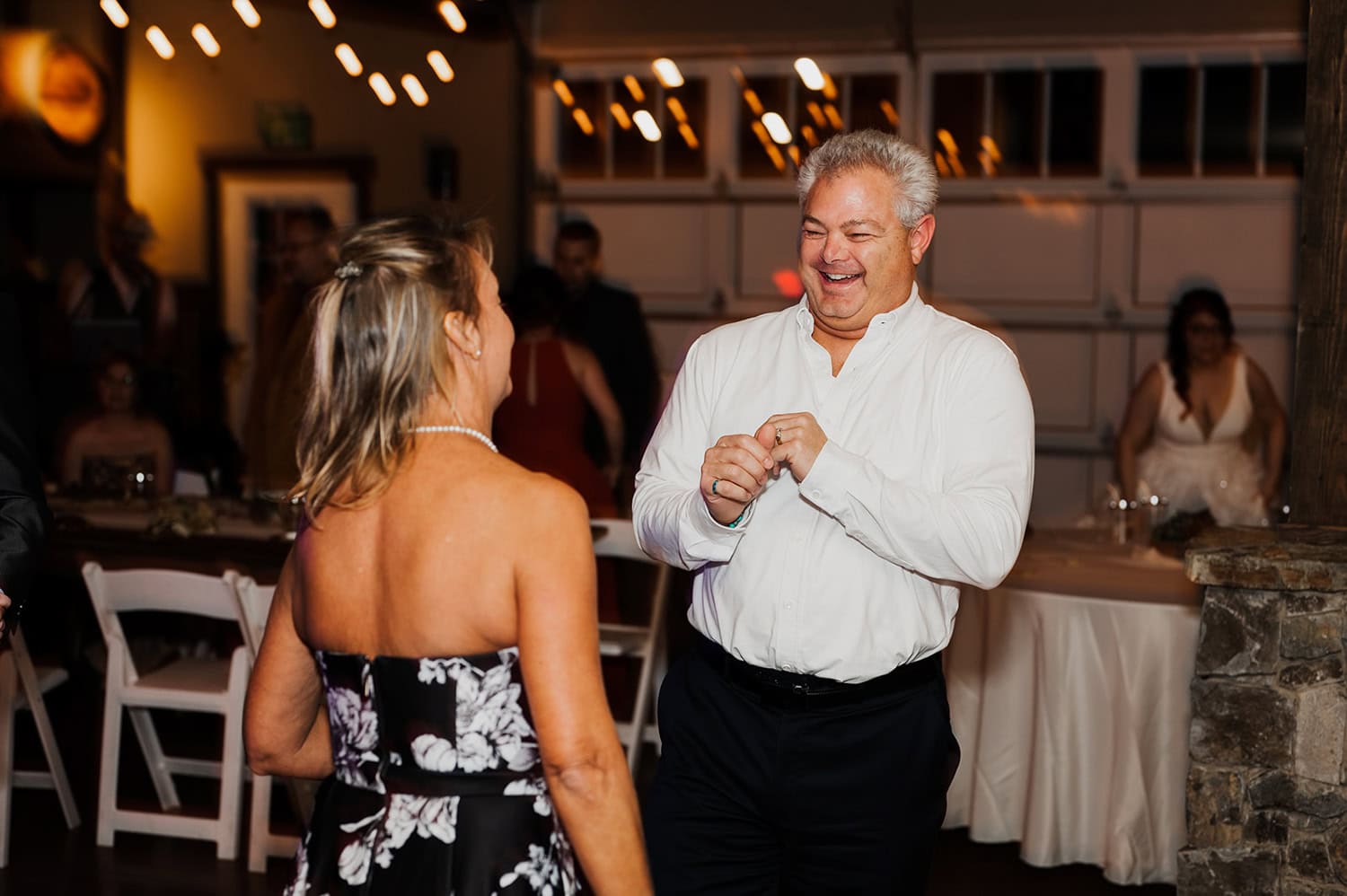 A man and woman dance together in a warmly lit indoor setting at Gorge Crest Vineyards during a wedding. Both are smiling, capturing the joyful atmosphere. Decor and guests can be seen blurred in the background, enhancing the lively environment.