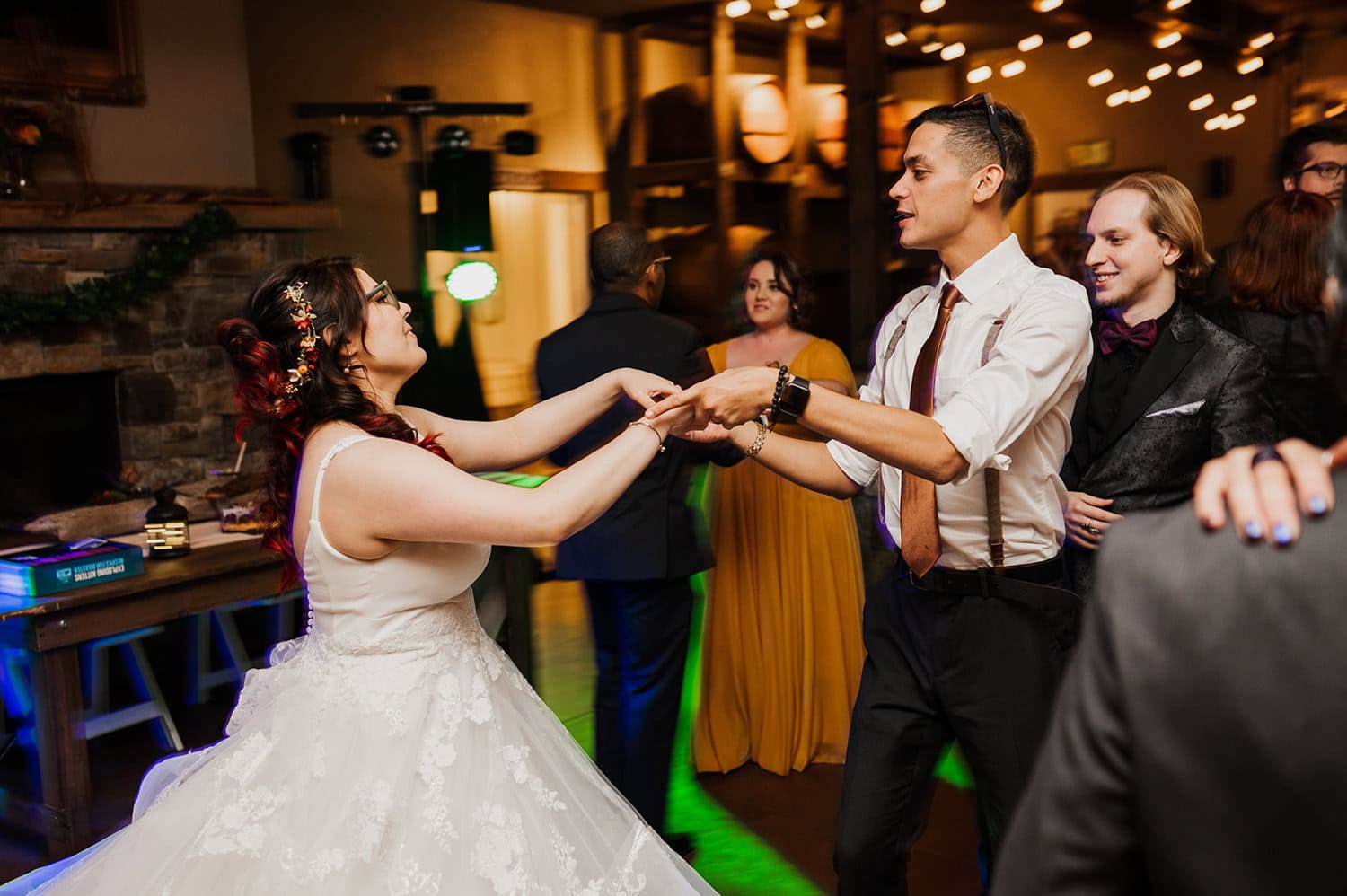 A bride and a man are dancing energetically together at an indoor wedding reception, with guests in the background smiling and clapping.