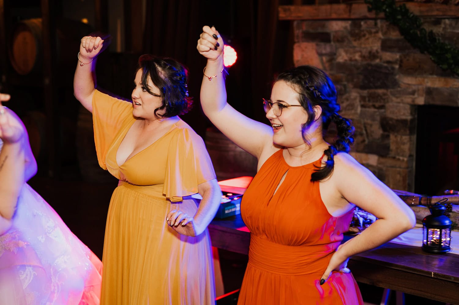 Two women in colorful dresses dance joyfully at an indoor party, with vibrant lighting casting bright hues against a stone wall backdrop.