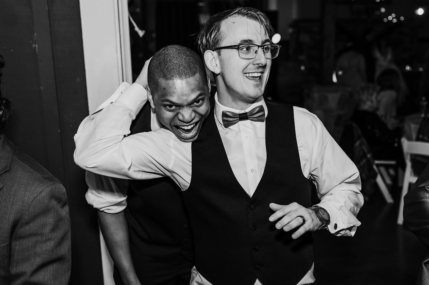 Two men in vests laughing and enjoying themselves together at an indoor event.