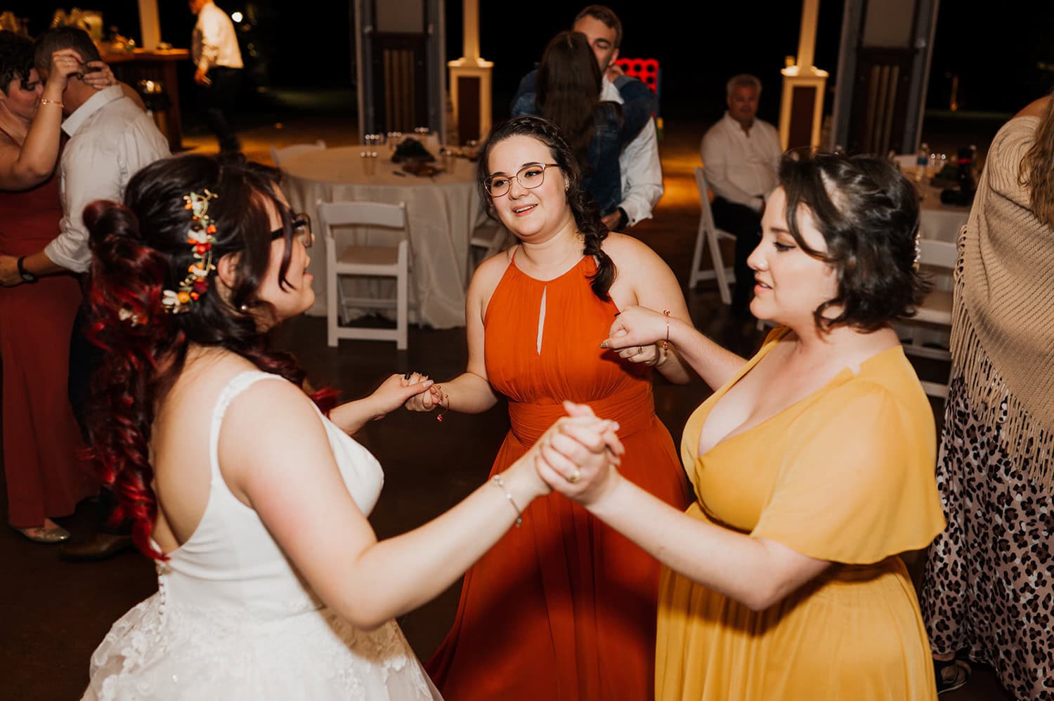 Three women in colorful dresses hold hands and dance joyfully at a lively indoor wedding event.