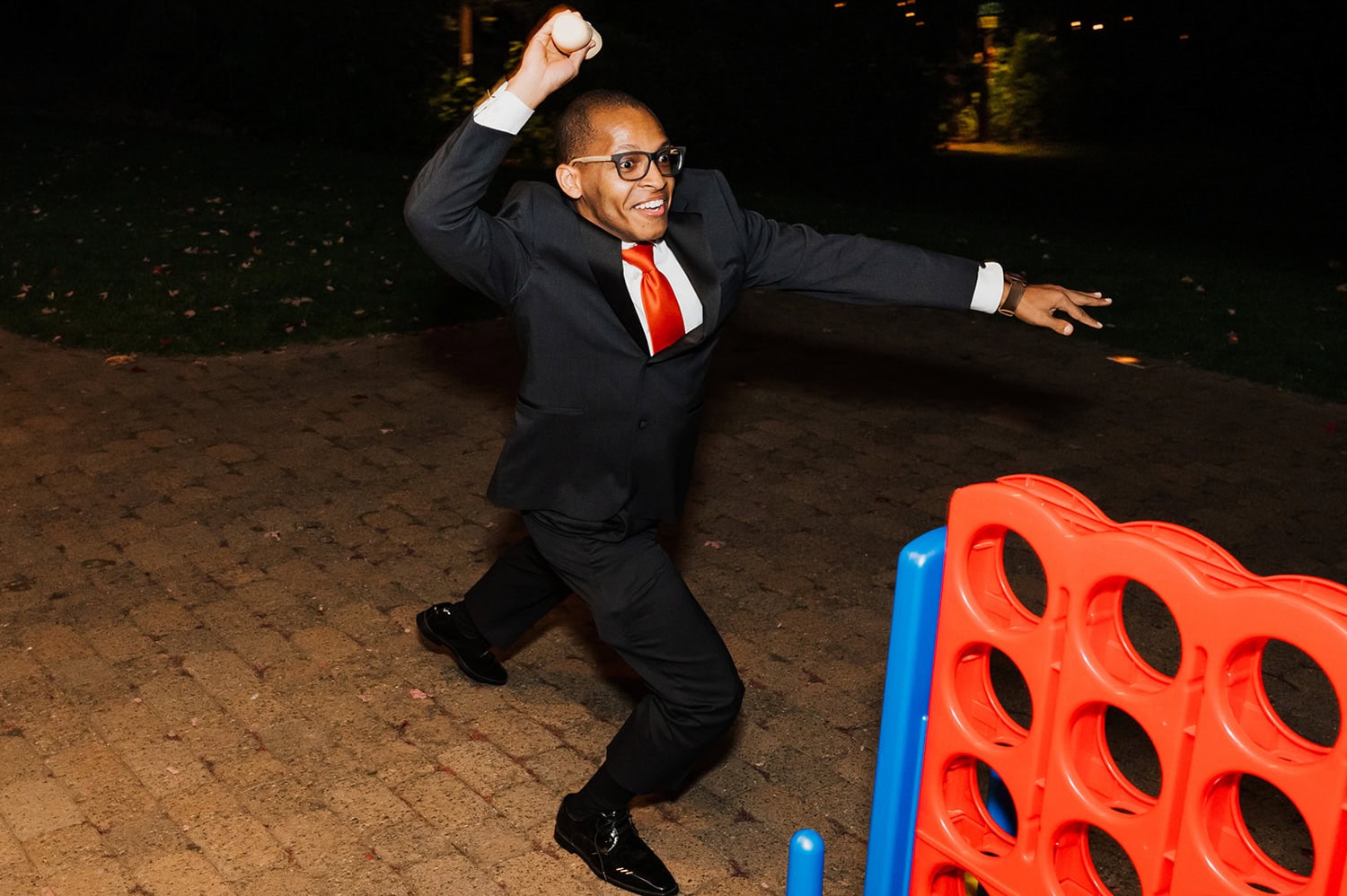 A man in a suit excitedly throws a ball at night near an oversized Connect Four game during a vibrant wedding at Gorge Crest Vineyards.
