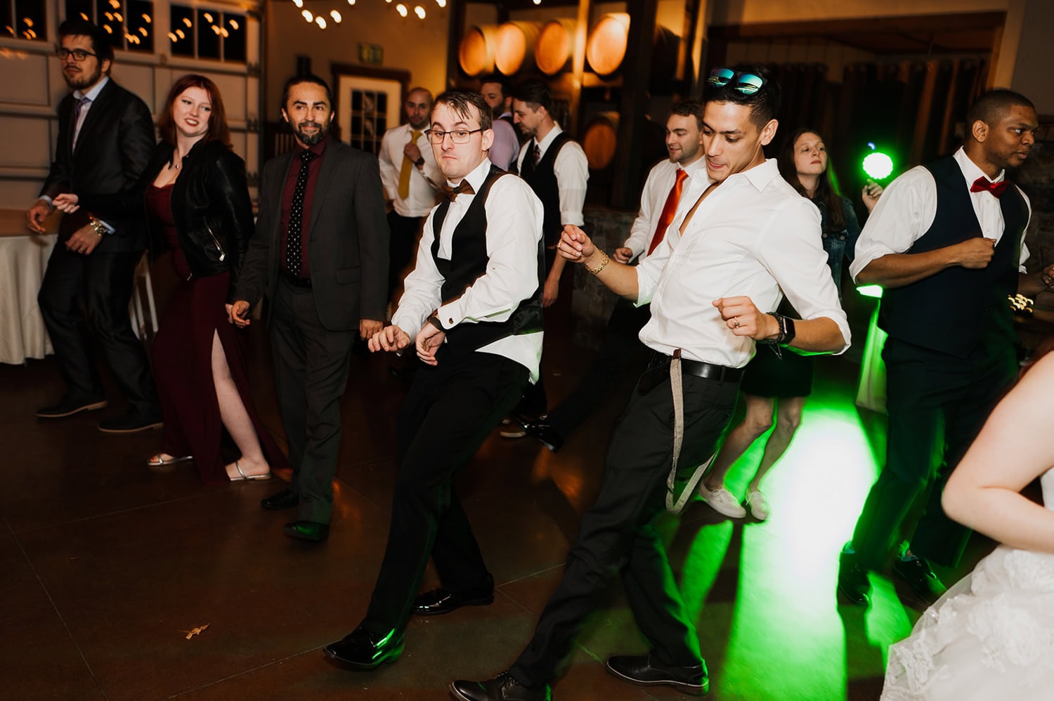 People dancing energetically at a wedding in Gorge Crest Vineyards, surrounded by green lights and oak barrels in a dimly lit room.