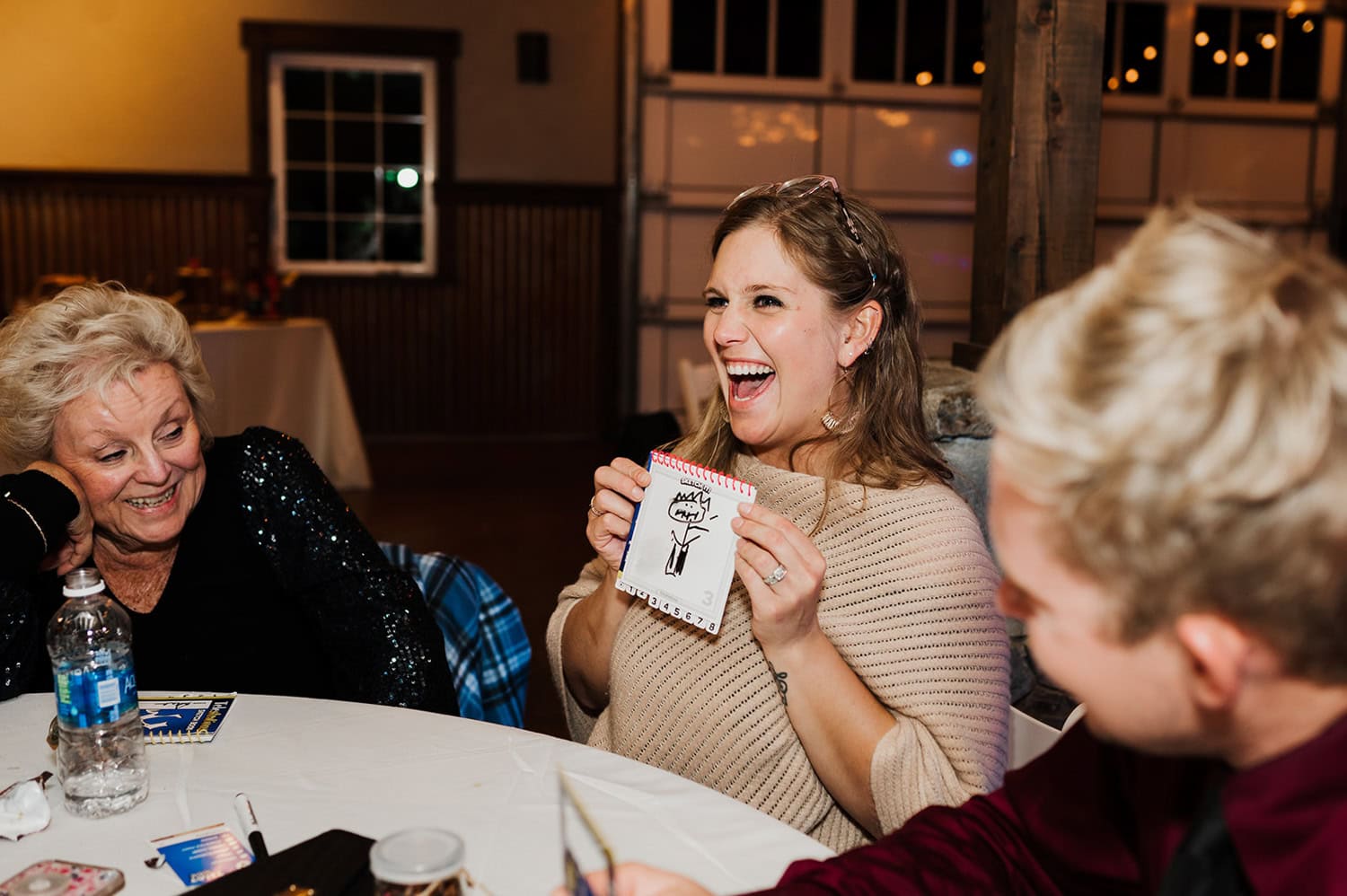 At Gorge Crest Vineyards, a smiling woman holds a doodle while seated at a table with two others, all laughing together during a wedding celebration.