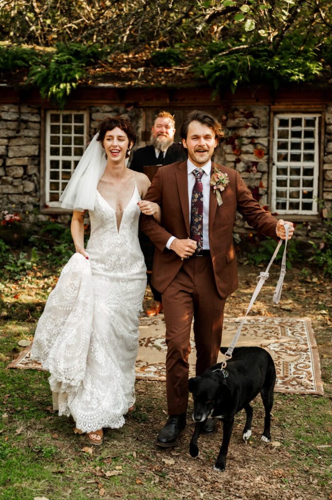 A smiling bride in a white dress and veil walks arm-in-arm with her cheerful new husband wearing a brown suit with their black dog on a leash up their wedding ceremony aisle on a lawn in a green lawn with a vintage rug on the ground in front of a stone hut.