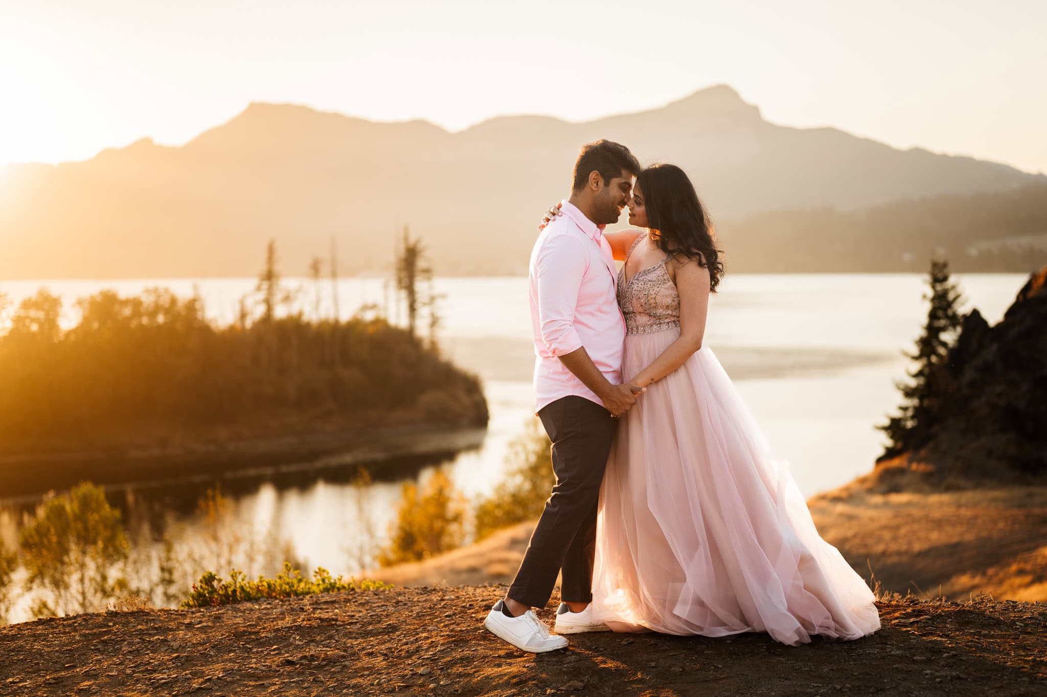 A couple on a cliffside embrace in front of a river during their Columbia River Gorge elopement, captured by Portland, Oregon elopement photographer Kyle Carnes. The bride wears a beautiful pink dress, and the groom wears a matching pink shirt.