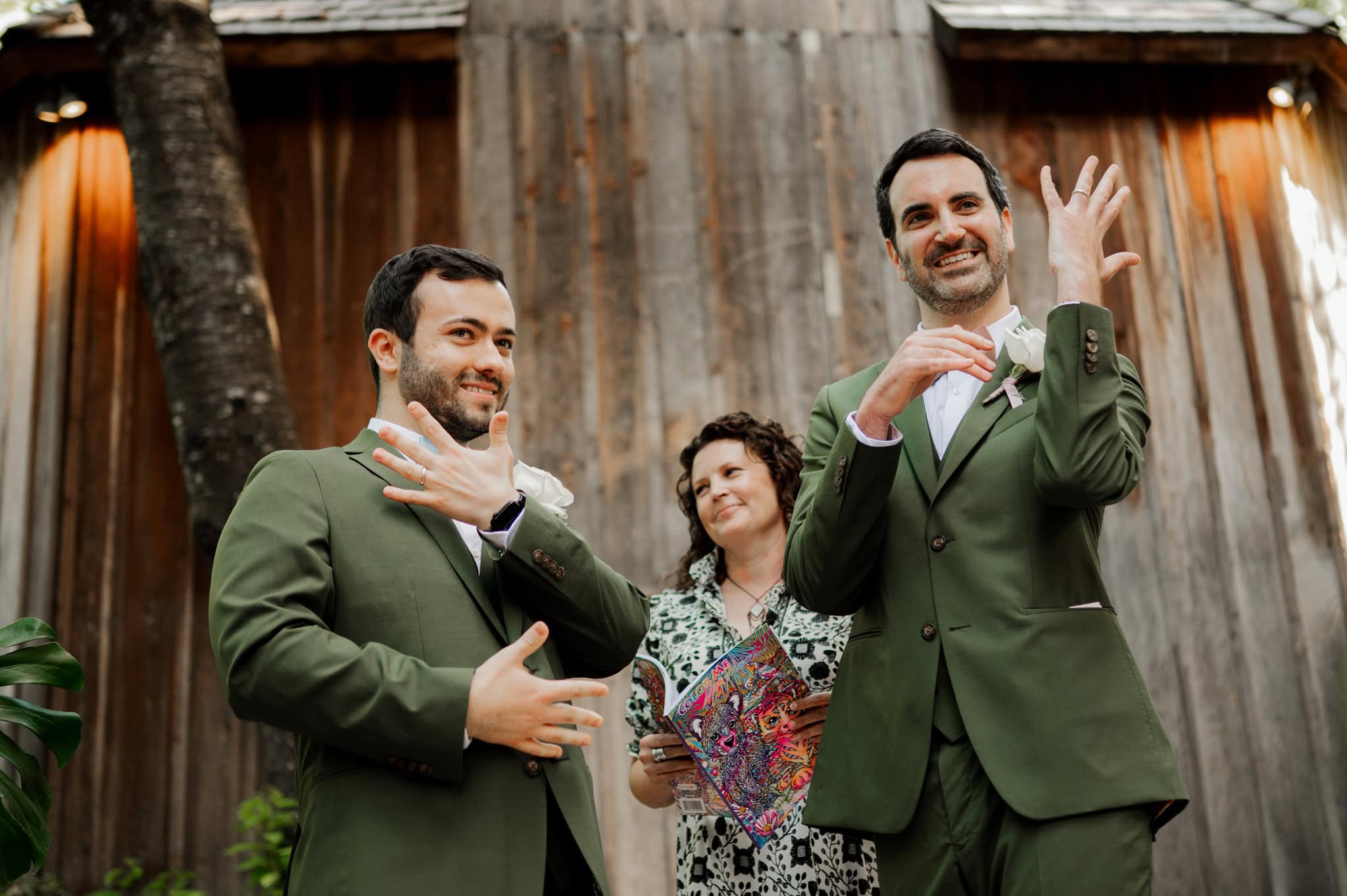 Two grooms in green suits joyfully showing rings and smiling, with a woman holding a colorful book behind them. They are standing in front of the rustic Octagonal Barn at McMenamins Cornelius Pass Roadhouse. Photo by Portland Oregon wedding and elopement photographer Kyle Carnes.