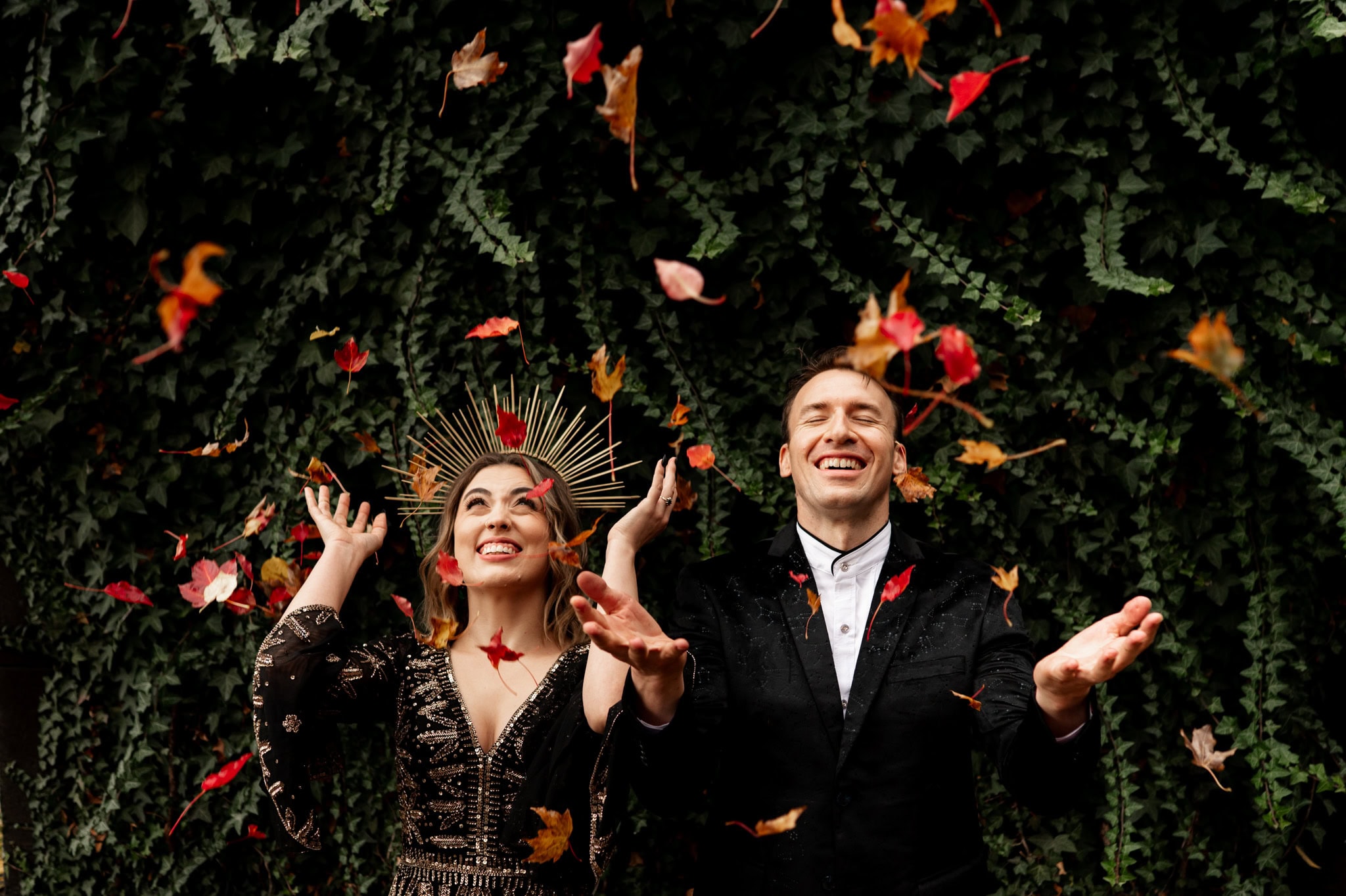 A joyful couple in black and gold formal attire throws autumn leaves into the air against a lush green ivy wall, photographed by Portland wedding and elopement photographer Kyle Carnes.