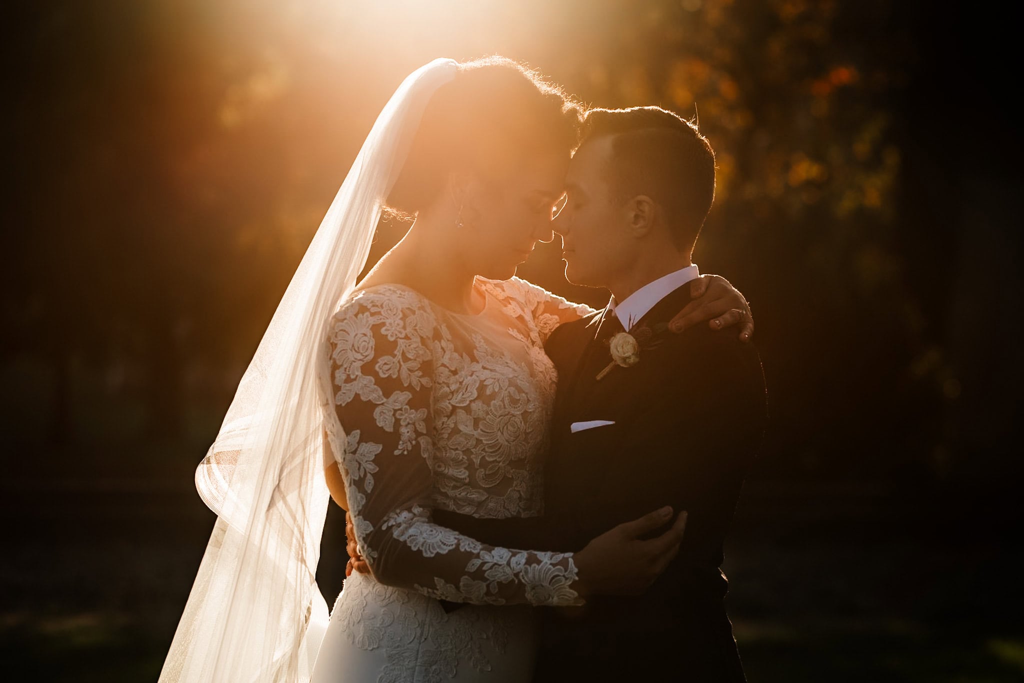 Bride and groom embrace at sunset, with soft, warm backlighting highlighting the brides lace dress and veil at Peninsula Park. Captured by Portland, Oregon wedding and elopement photographer Kyle Carnes.