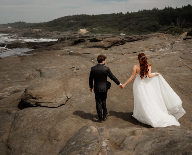 The bride and groom walk hand in hand across a rocky coastal landscape with a cloudy sky above, taking wedding photos at the Yachats Inn.