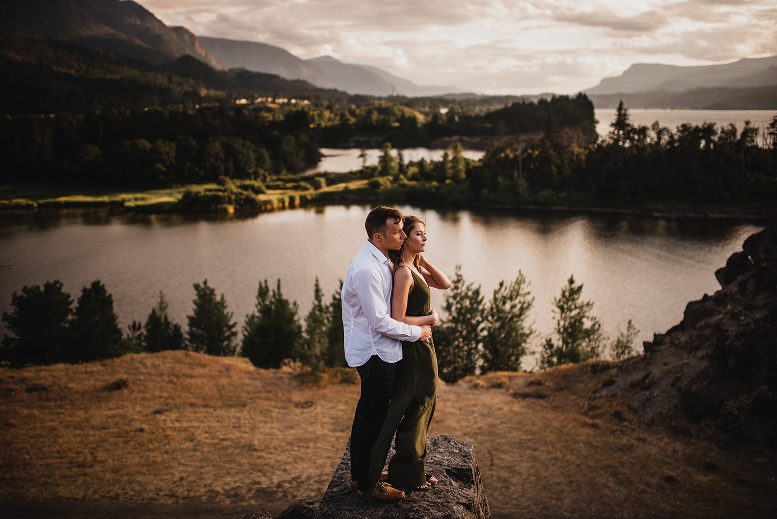 An engaged couple stands together on a large rocky outcropping, gazing out over the Columbia River surrounded by mountains with the sky glowing in warm sunset colors.
