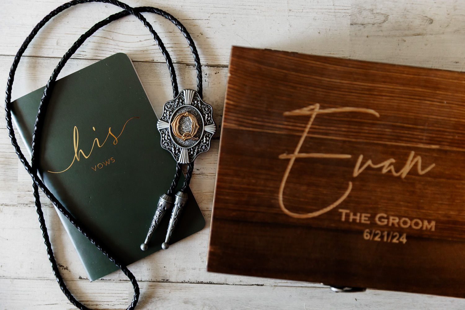 A grooms vows book, a bolo tie, and a personalized wooden box are arranged on a white wooden surface.