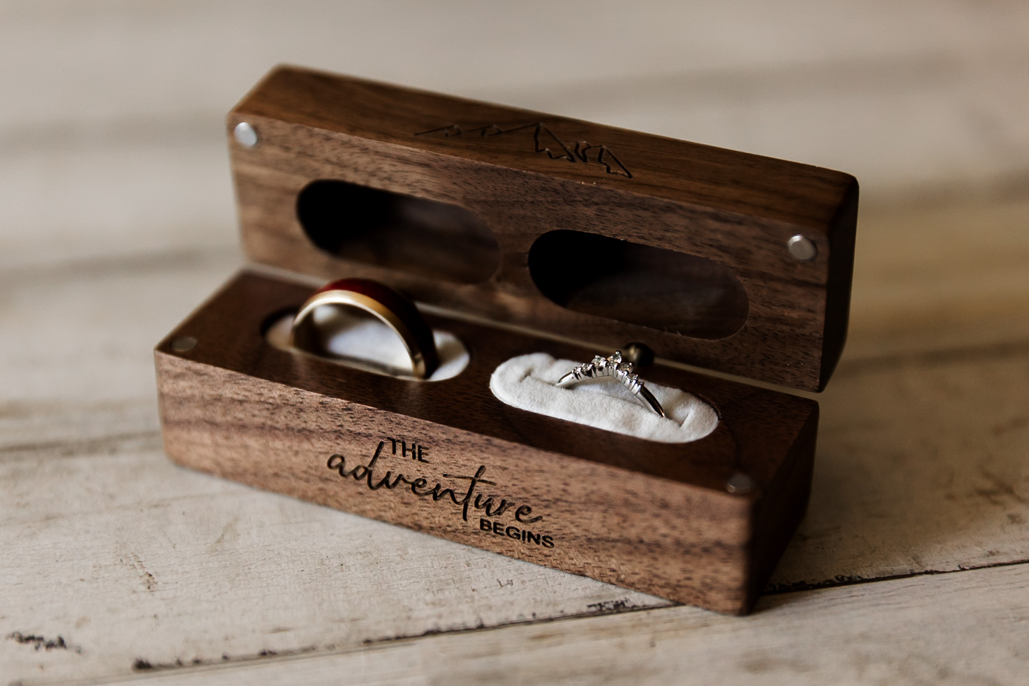 In a wooden box with the inscription The Adventure Begins, a gold band and diamond ring are placed on white fabric, signifying preparation for a wedding at the Hoyt Arboretum Redwood Deck.