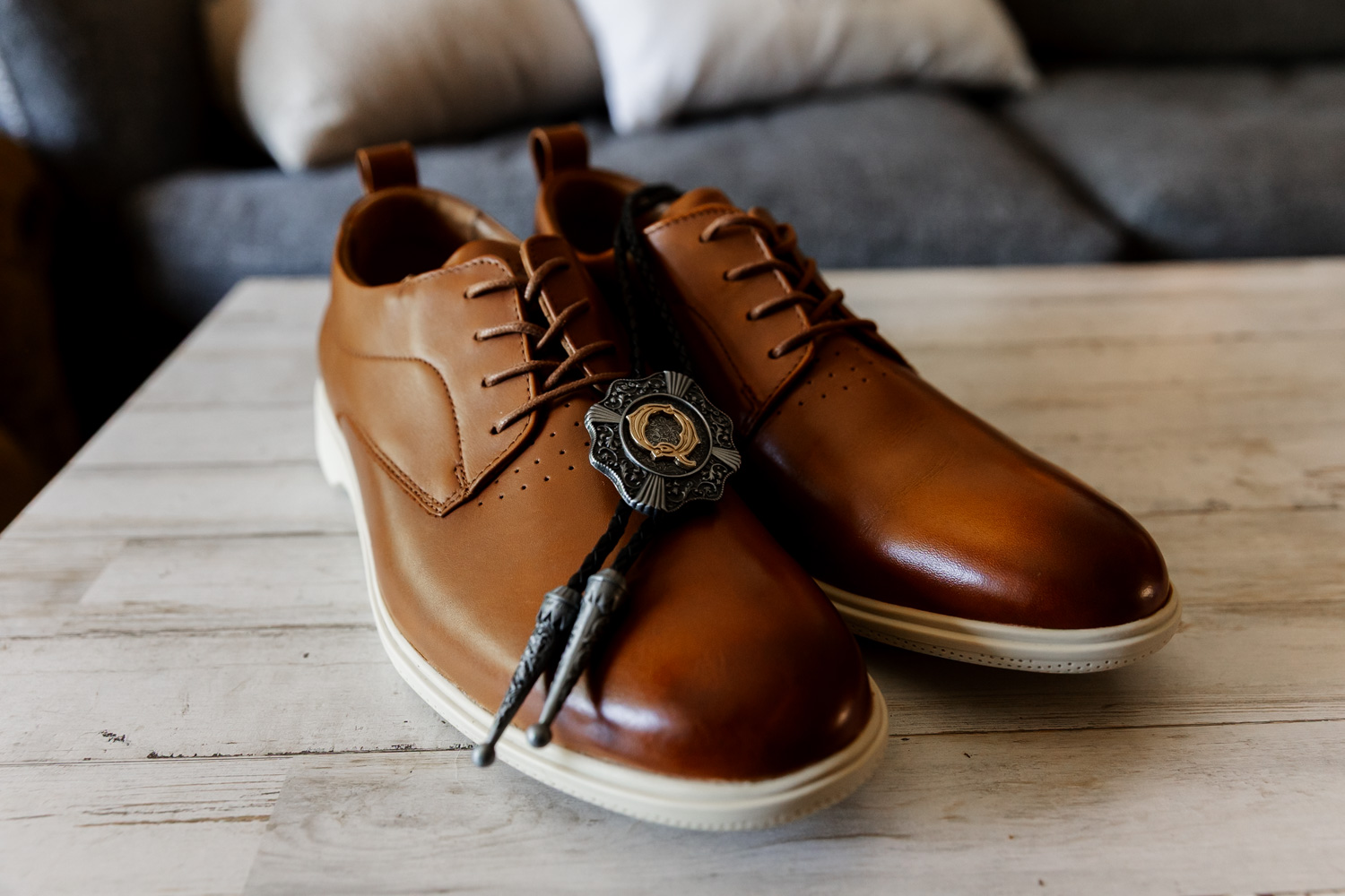 A pair of brown leather shoes on a wooden table with a decorative bolo tie placed on top.