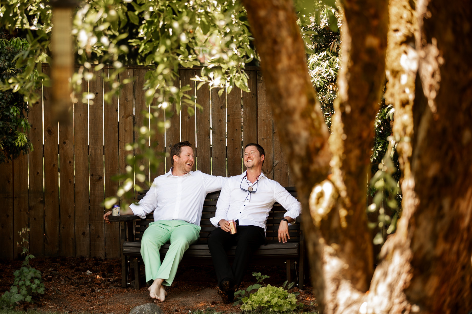Two men sit laughing on a bench in a garden, preparing for a wedding at the Hoyt Arboretum Redwood Deck, surrounded by trees and a wooden fence.