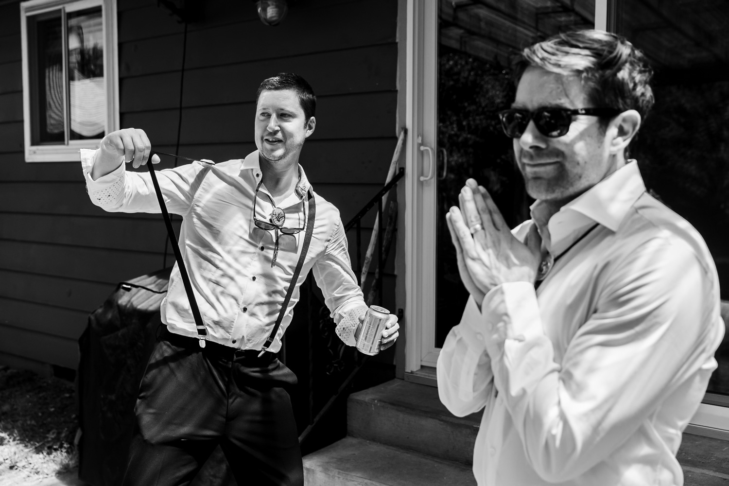 Two men are preparing for a wedding at the Hoyt Arboretum Redwood Deck. They are dressed in formal attire. One man is adjusting his suspenders, while the other stands beside him, smiling and clasping his hands together.