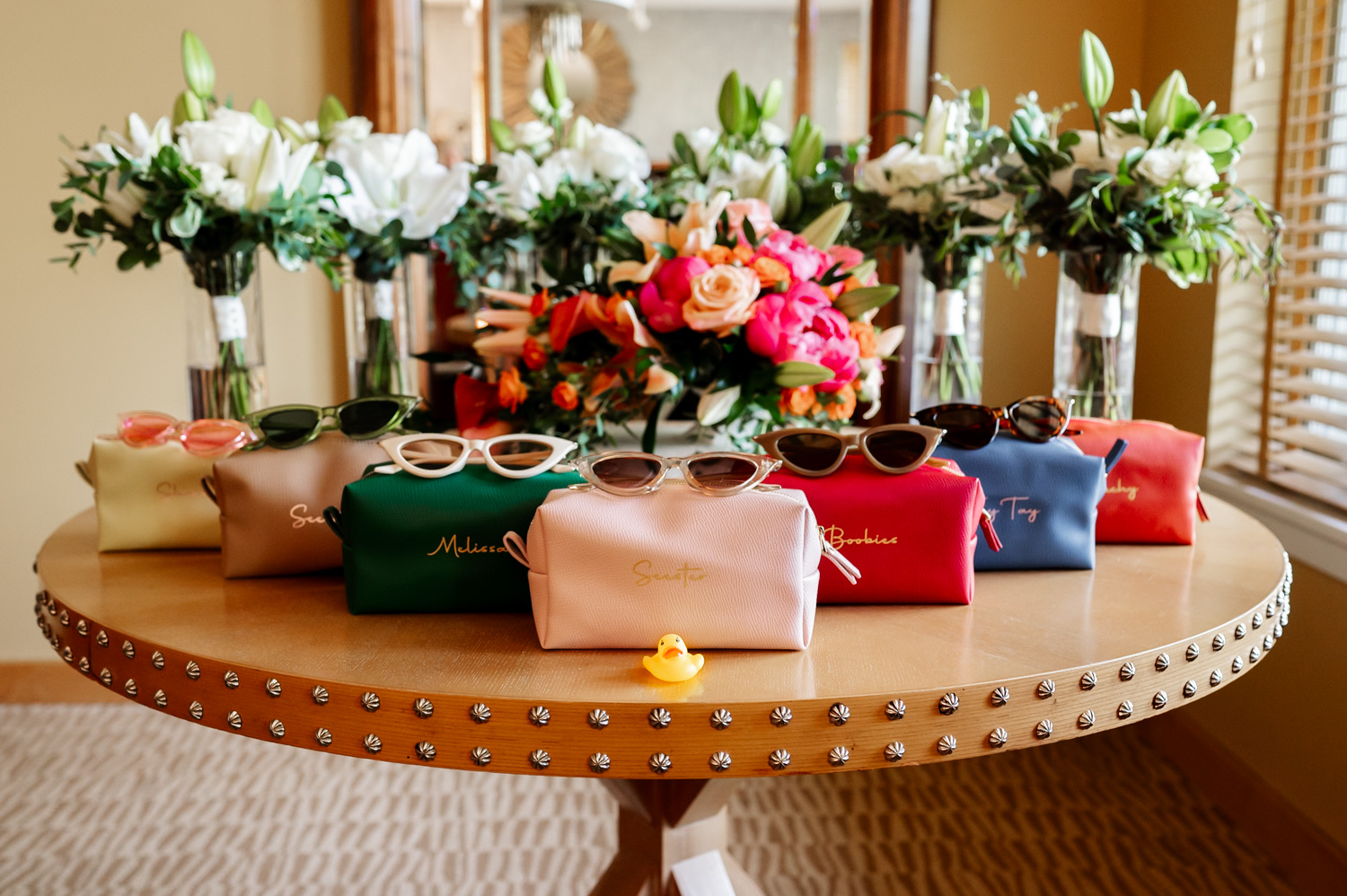 A collection of cheerful sunglasses and personalized bags lies scattered across a table, with vibrant flower bouquets adding bursts of color. A small yellow rubber duck playfully perches among the items, all set against the backdrop of preparations for a wedding at Hoyt Arboretum Redwood Deck.