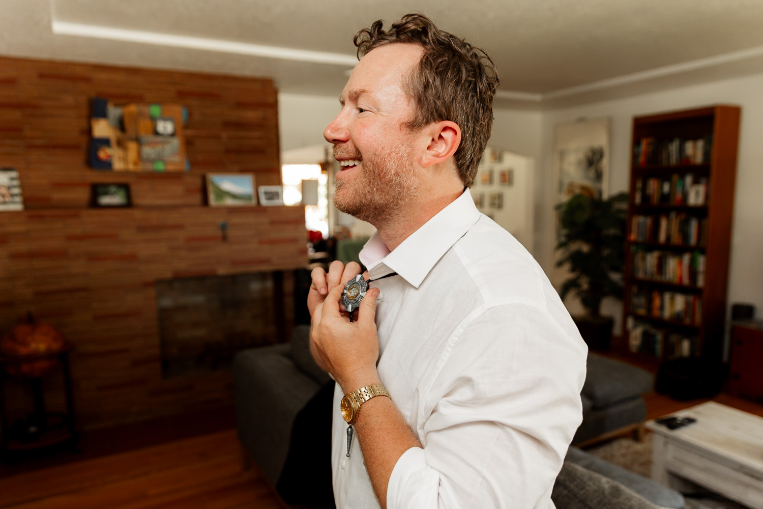 A smiling man in a white shirt buttoning up, preparing for an event, stands in a cozy living room with bookshelves and a fireplace.