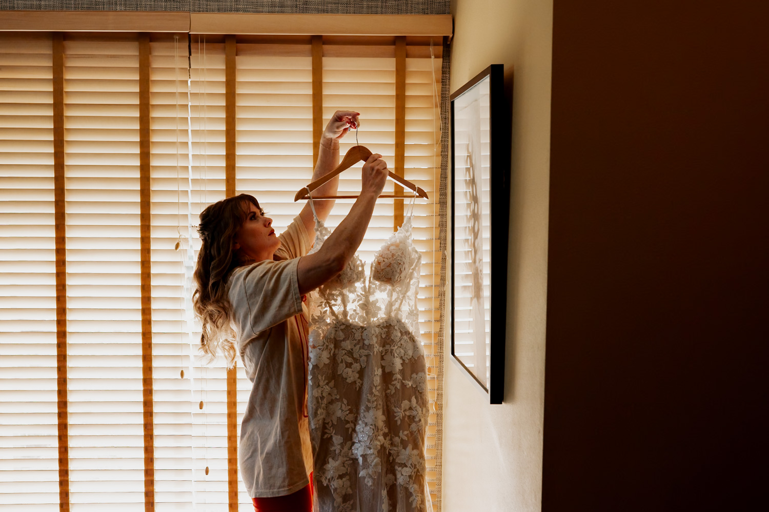 A woman holds a floral wedding dress on a hanger near window blinds, preparing for a wedding at the Hoyt Arboretum Redwood Deck.