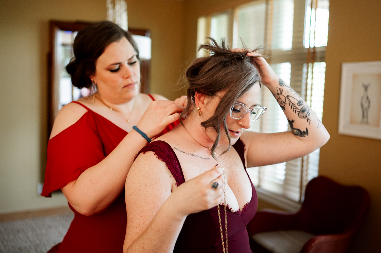 In a warmly lit room, two women in red dresses stand close together as one adjusts the necklace of the other, sharing a tender moment before attending a wedding at Hoyt Arboretum Redwood Deck.