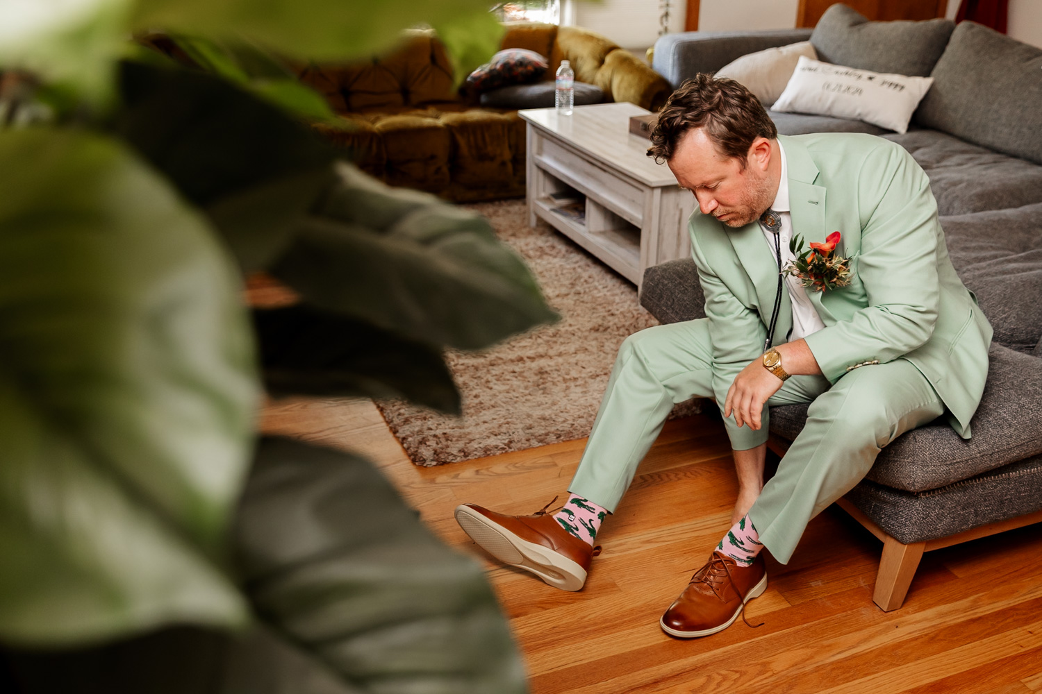 A man in a mint green suit sits on a couch, adjusting his colorful socks and wearing brown shoes. He is preparing for a wedding at the Hoyt Arboretum Redwood Deck. The setting includes leafy plants and elegant furniture.