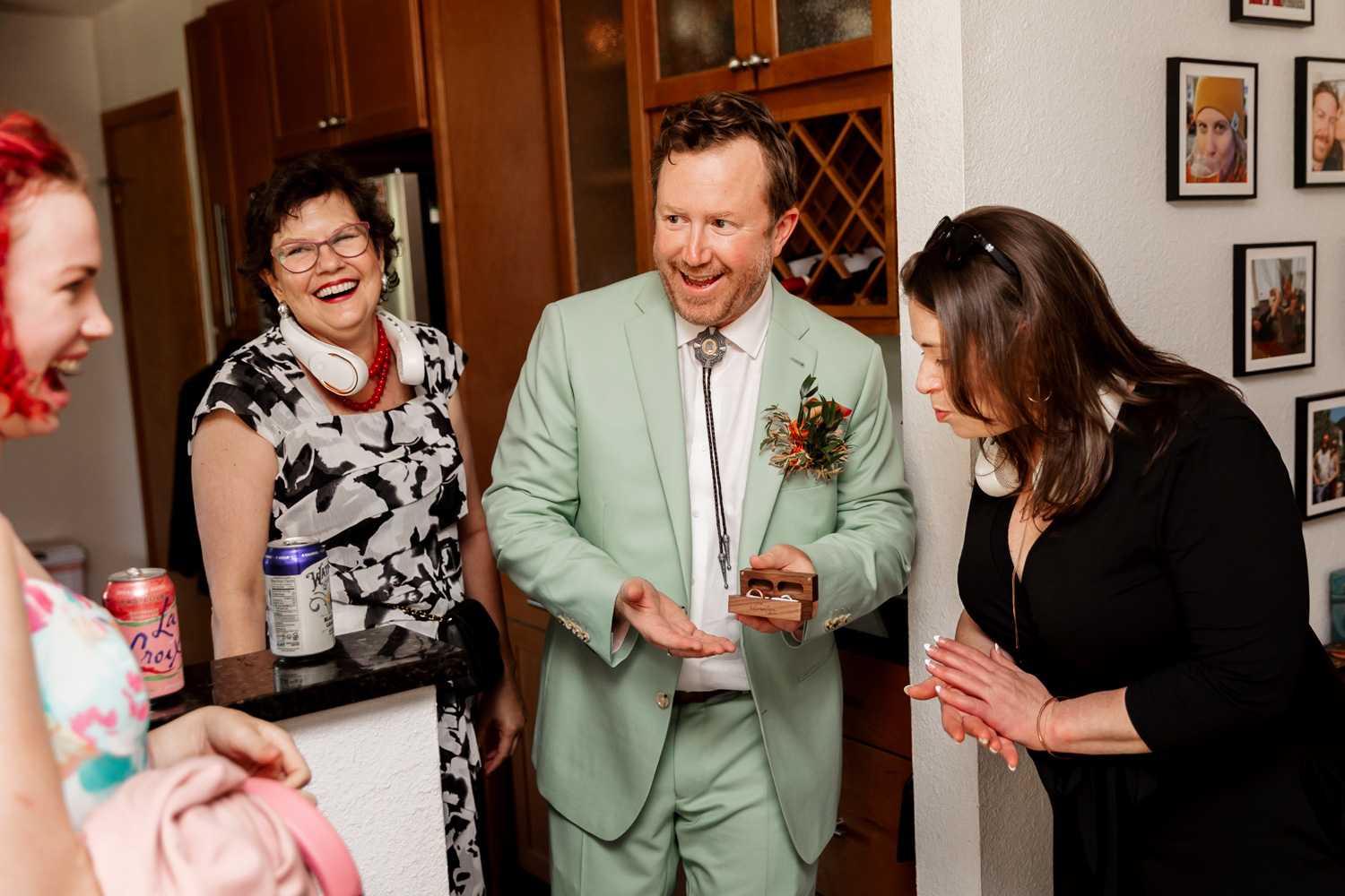A smiling man in a light green suit holds up a small box to three amused people in a room as they prepare for a wedding at the Hoyt Arboretum Redwood Deck.